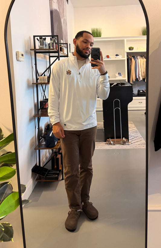 A man takes a mirror selfie in a boutique showing a white long-sleeve quarter-zip shirt with a small LSU Vintage Tiger embroidered logo on the left chest, paired with brown pants and brown shoes.