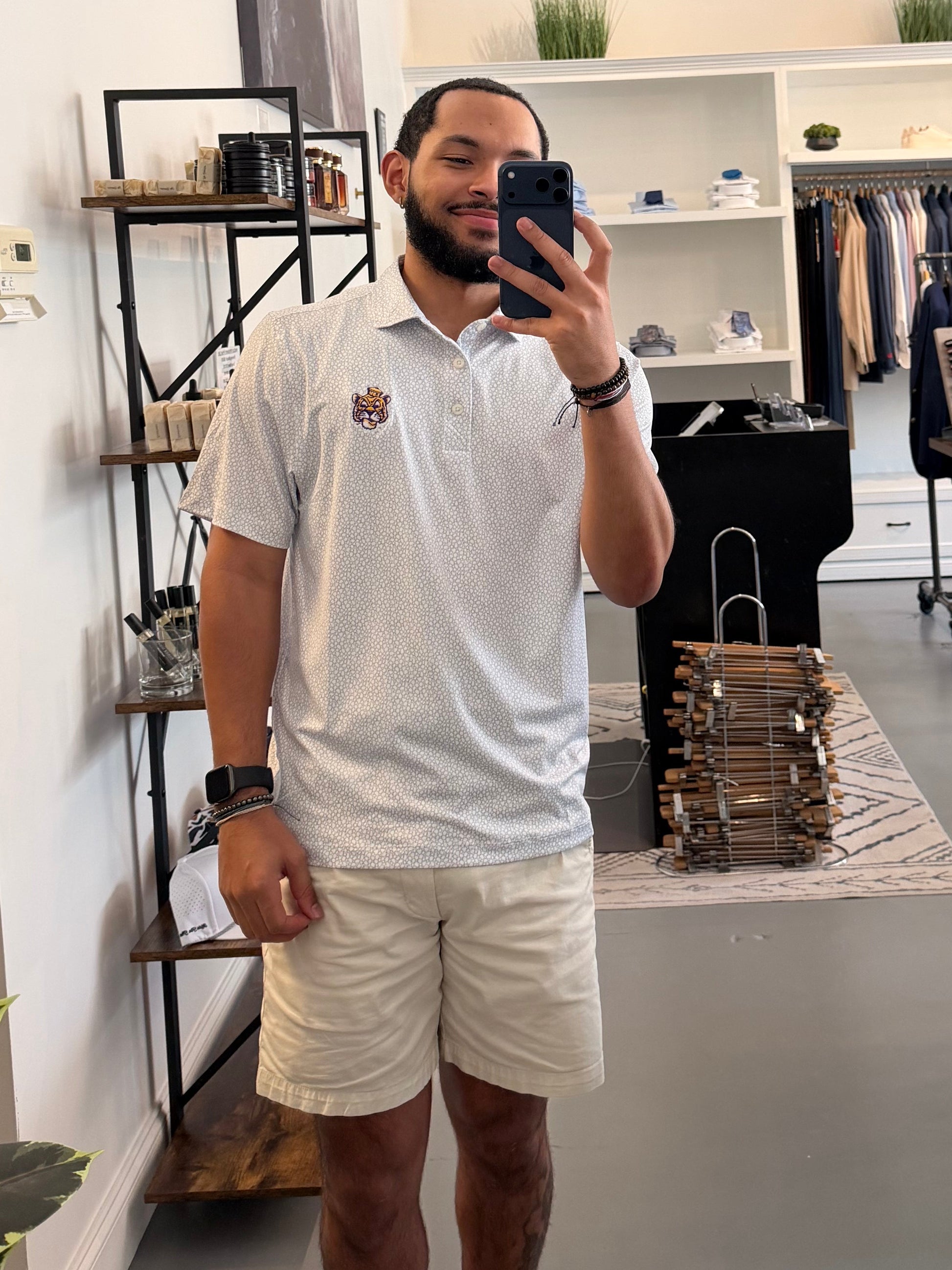 A man takes a mirror selfie in a clothing boutique, wearing a LSU Sailor Mike white pebble polo shirt with a small LSU Sailor Mike the tiger embroidered logo on the chest and beige shorts. The boutique features a large arched floor mirror, clothing racks, and shelves in the background.