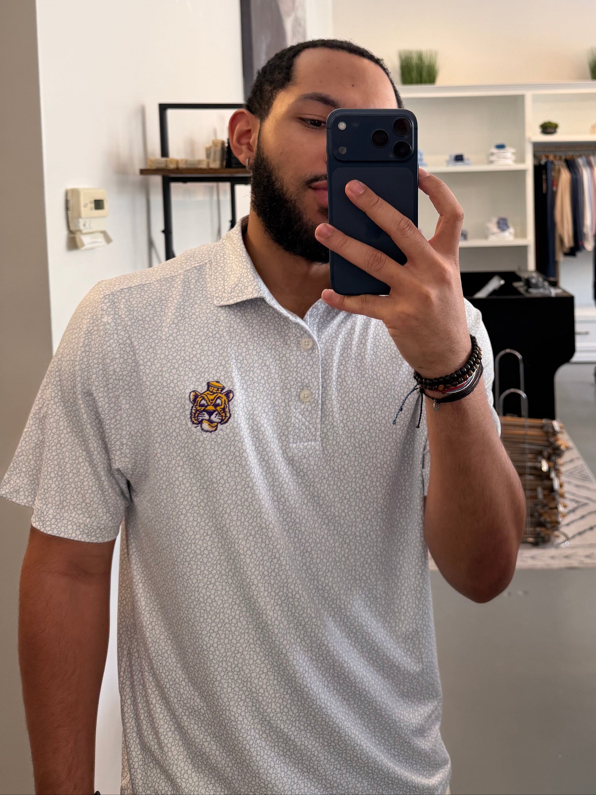 A man takes a mirror selfie in a clothing boutique, wearing a LSU Sailor Mike white pebble polo shirt with a small LSU Sailor Mike the tiger embroidered logo on the chest and beige shorts. The boutique features a large arched floor mirror, clothing racks, and shelves in the background.