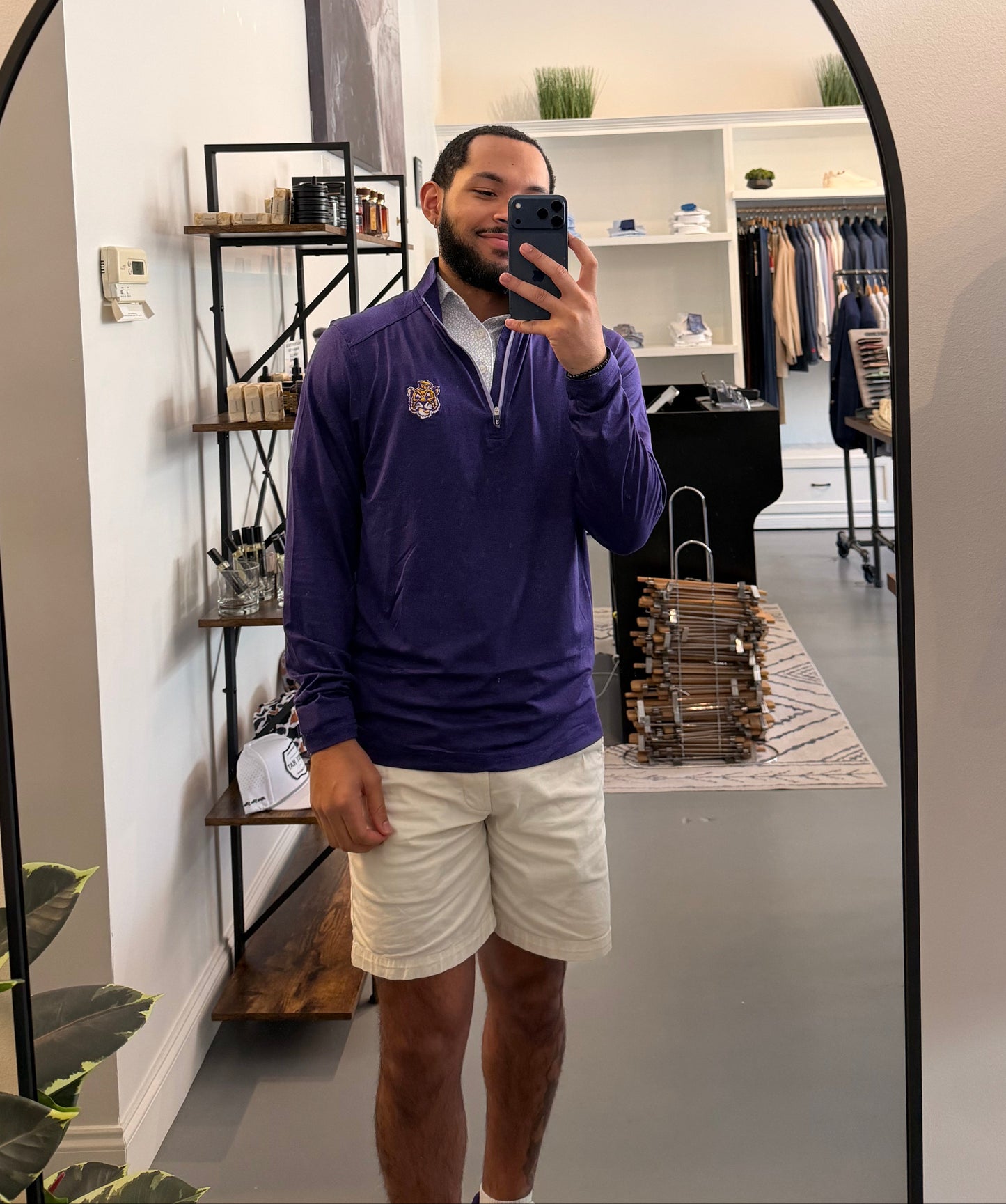 A man takes a mirror selfie in a boutique while wearing a LSU Tiger Sailor Mike purple zip-up hoodie with a white collar and a small Sailor Mike Lsu Tiger crest logo on the chest, paired with white short sleeve Lsu Tiger Pebble print polo. 