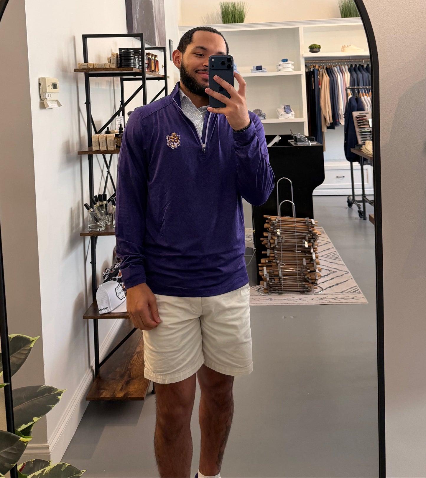 A man takes a mirror selfie in a boutique while wearing a LSU Tiger Sailor Mike purple zip-up hoodie with a white collar and a small Sailor Mike Lsu Tiger crest logo on the chest, paired with white short sleeve Lsu Tiger Pebble print polo. 