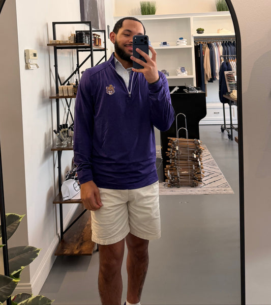 A man takes a mirror selfie in a boutique while wearing a LSU Tiger Sailor Mike purple zip-up hoodie with a white collar and a small Sailor Mike Lsu Tiger crest logo on the chest, paired with white short sleeve Lsu Tiger Pebble print polo. 
