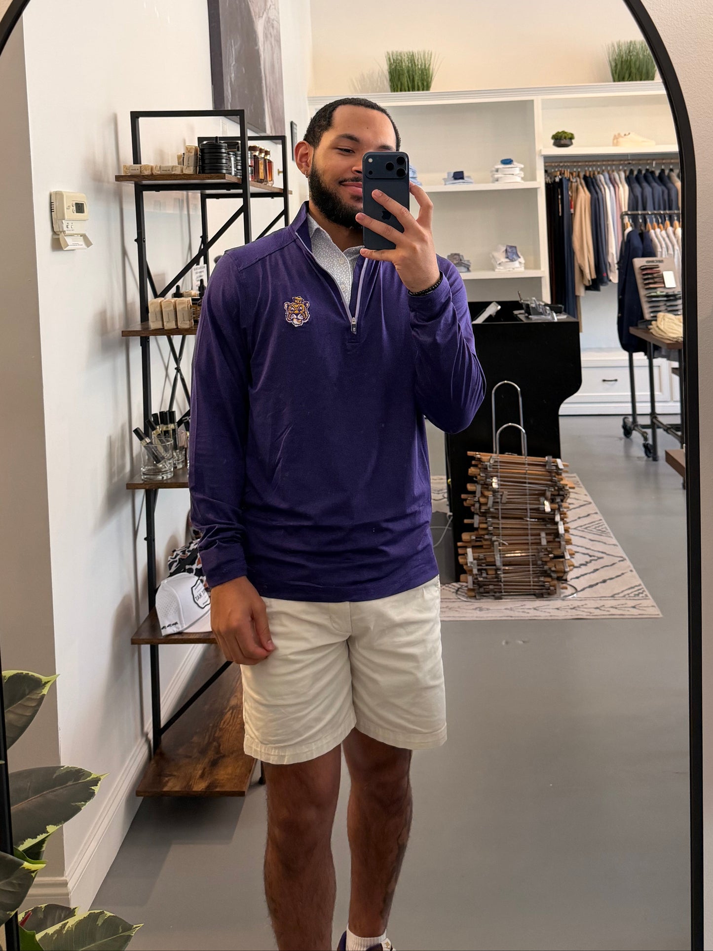 A man takes a mirror selfie in a boutique while wearing a LSU Tiger Sailor Mike purple zip-up hoodie with a white collar and a small Sailor Mike Lsu Tiger crest logo on the chest, paired with white short sleeve Lsu Tiger Pebble print polo. 