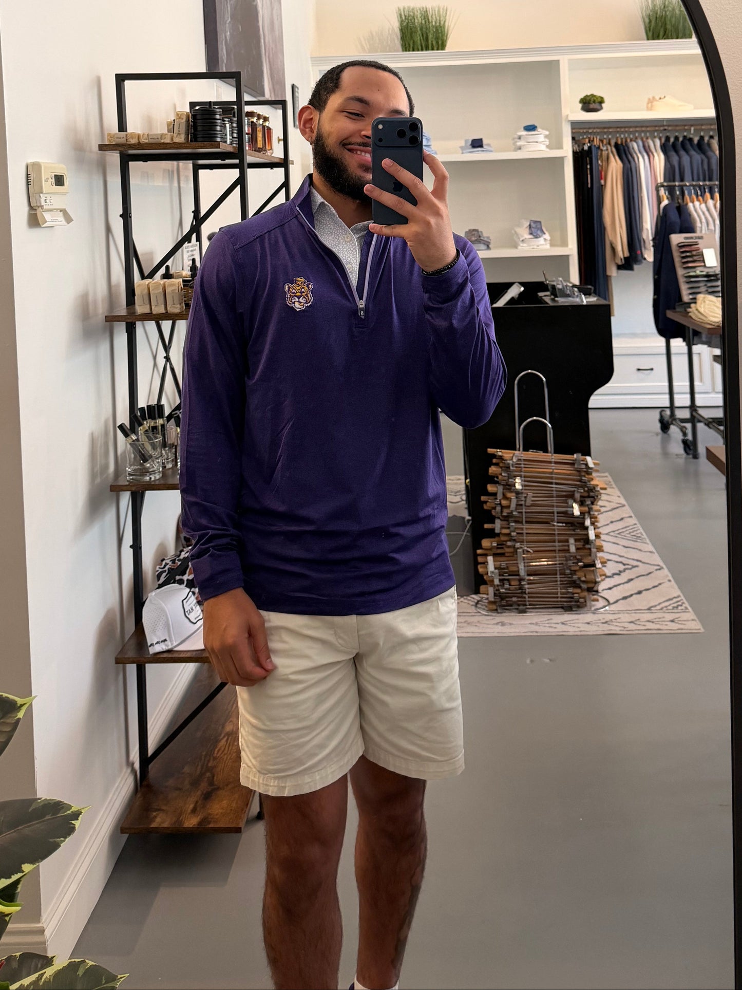 A man takes a mirror selfie in a boutique while wearing a LSU Tiger Sailor Mike purple zip-up hoodie with a white collar and a small Sailor Mike Lsu Tiger crest logo on the chest, paired with white short sleeve Lsu Tiger Pebble print polo. 
