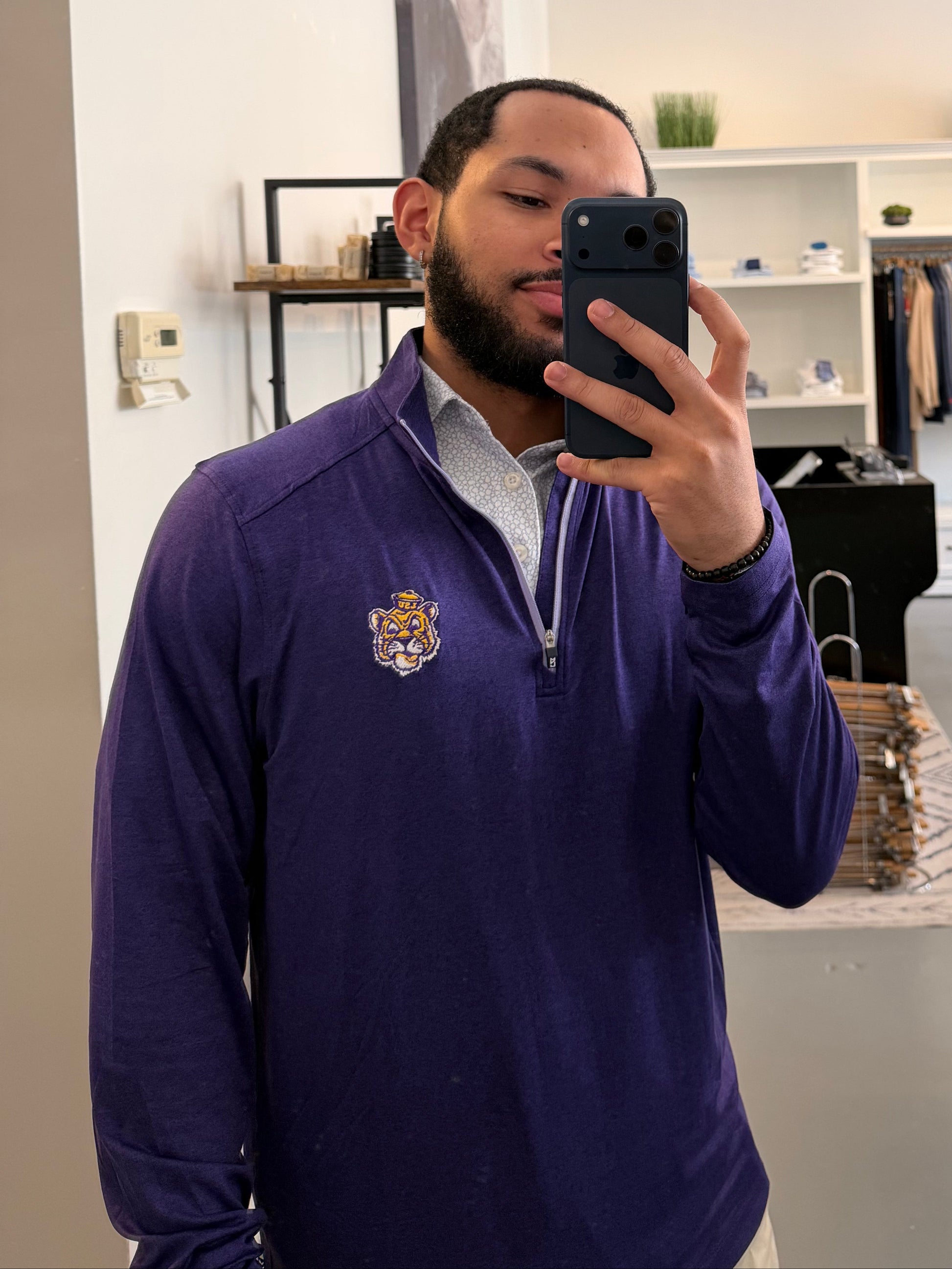 A man takes a mirror selfie in a boutique while wearing a LSU Tiger Sailor Mike purple zip-up hoodie with a white collar and a small Sailor Mike Lsu Tiger crest logo on the chest, paired with white short sleeve Lsu Tiger Pebble print polo. 
