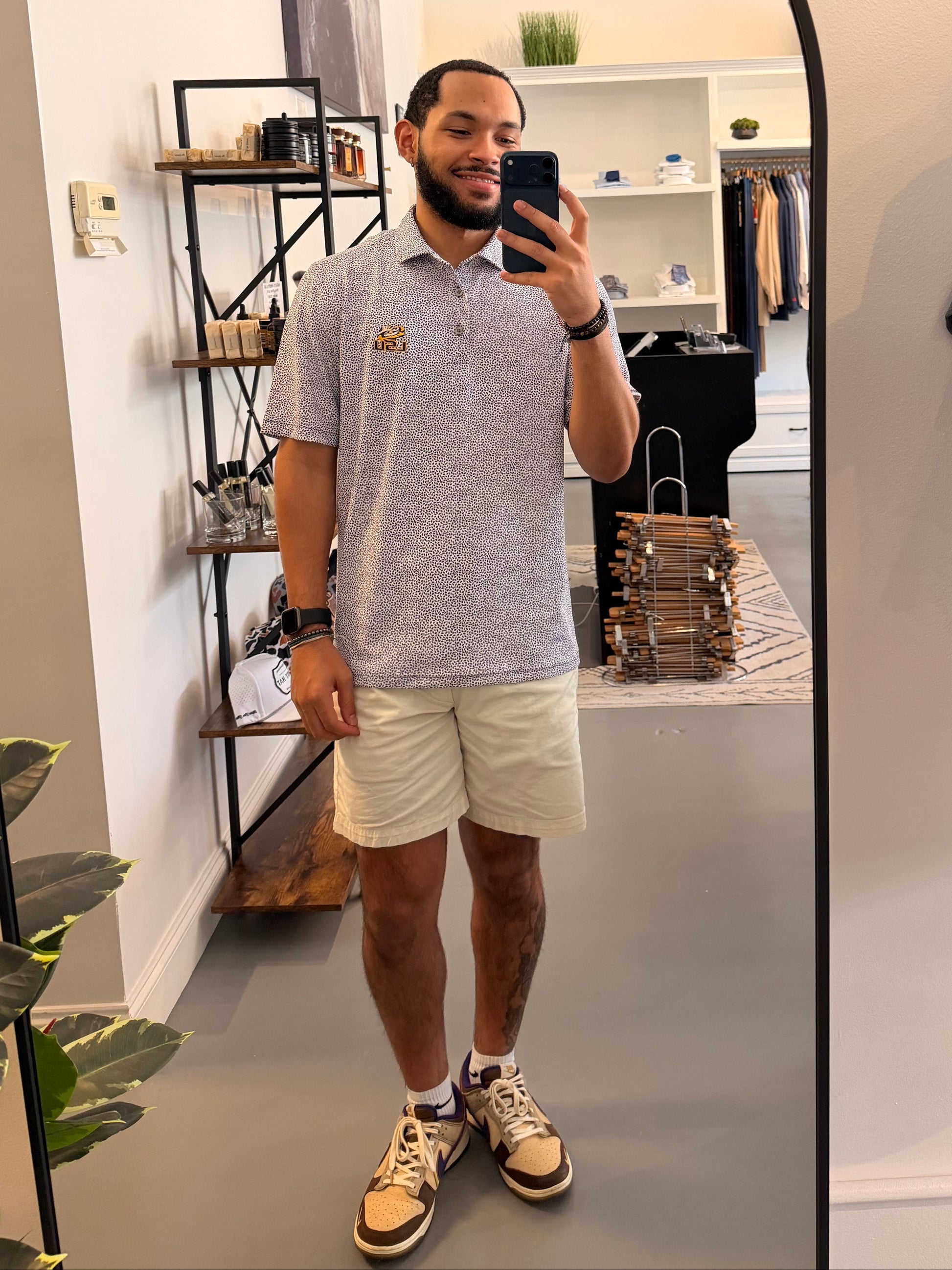 A man takes a mirror selfie in a boutique showing a botanical-floral college purple Tiger Eye Lsu Tiger polo shirt with a small LSU Tiger embroidered logo on the chest, paired with beige shorts and sneakers.