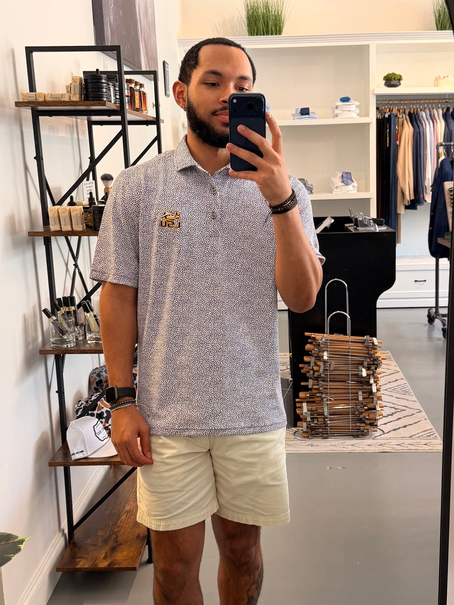 A man takes a mirror selfie in a boutique showing a botanical-floral college purple Tiger Eye Lsu Tiger polo shirt with a small LSU Tiger embroidered logo on the chest, paired with beige shorts and sneakers.