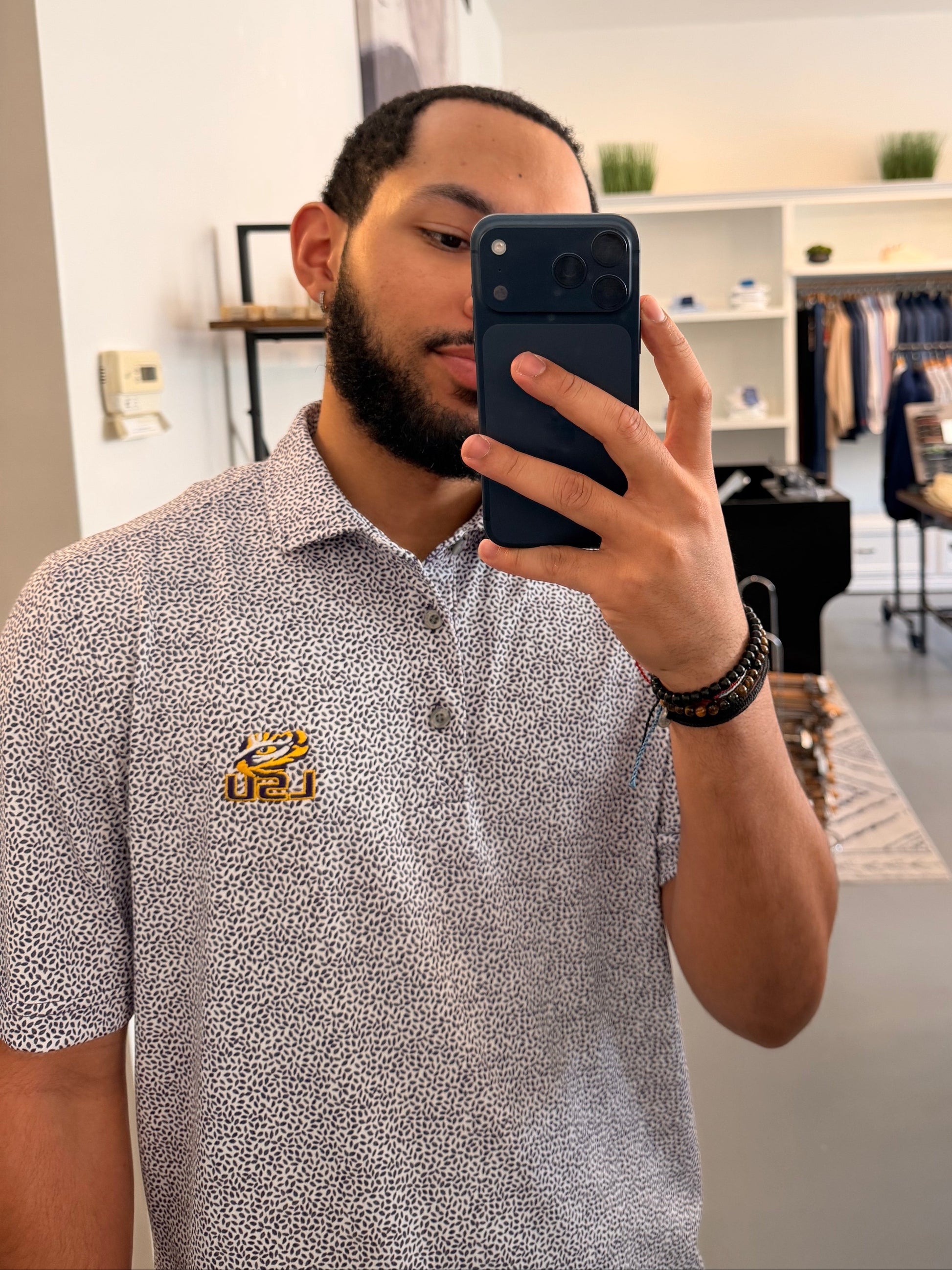 A man takes a mirror selfie in a boutique showing a botanical-floral college purple Tiger Eye Lsu Tiger polo shirt with a small LSU Tiger embroidered logo on the chest, paired with beige shorts and sneakers.