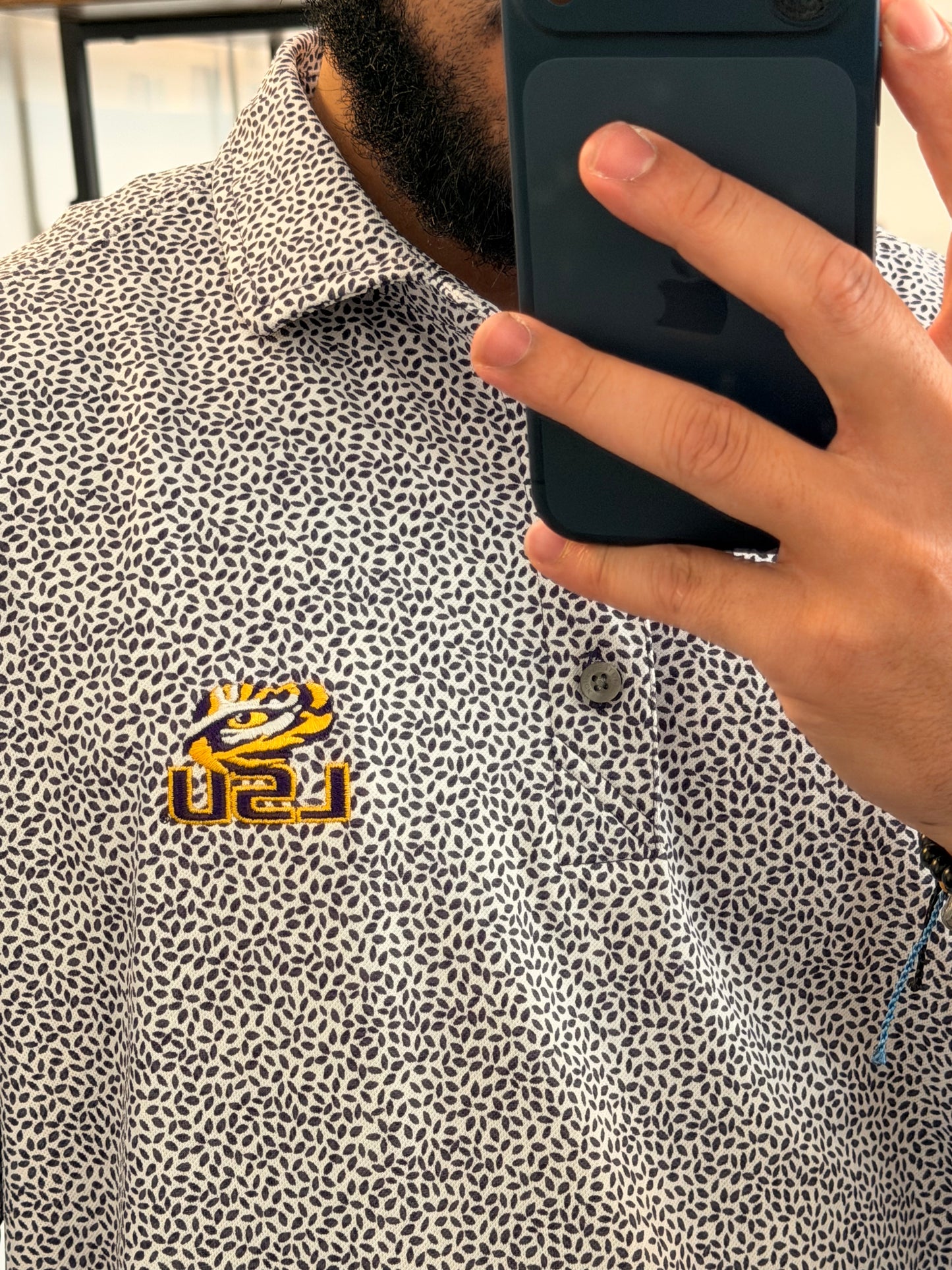 A man takes a mirror selfie in a boutique showing a botanical-floral college purple Tiger Eye Lsu Tiger polo shirt with a small LSU Tiger embroidered logo on the chest, paired with beige shorts and sneakers.