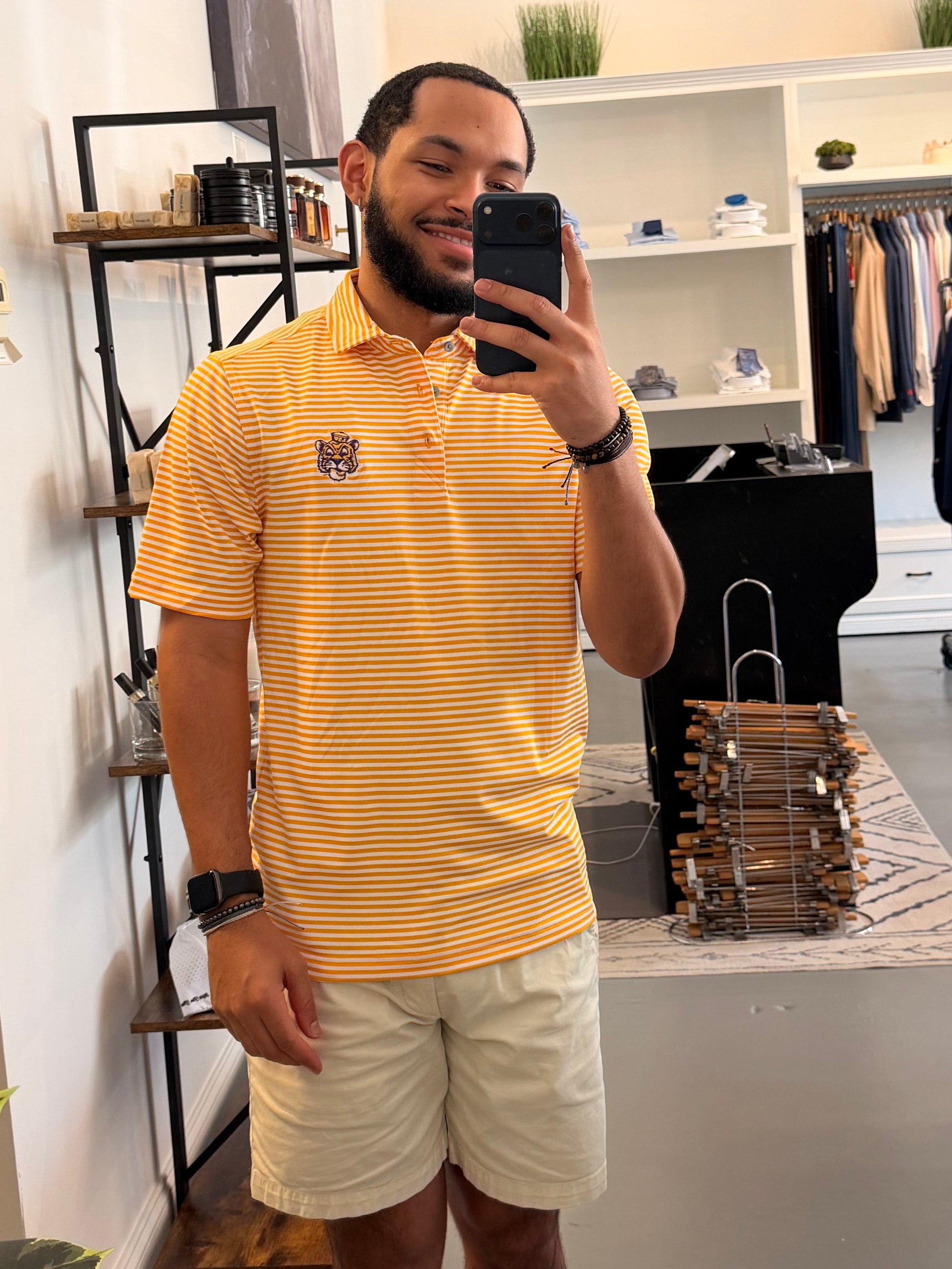 A man takes a mirror selfie in a clothing boutique wearing a LSU Sailor Mike the Tiger college gold and white striped polo shirt, beige shorts, and sneakers. The boutique features a large arched floor mirror with a black frame, white walls, and clothing racks in the background.