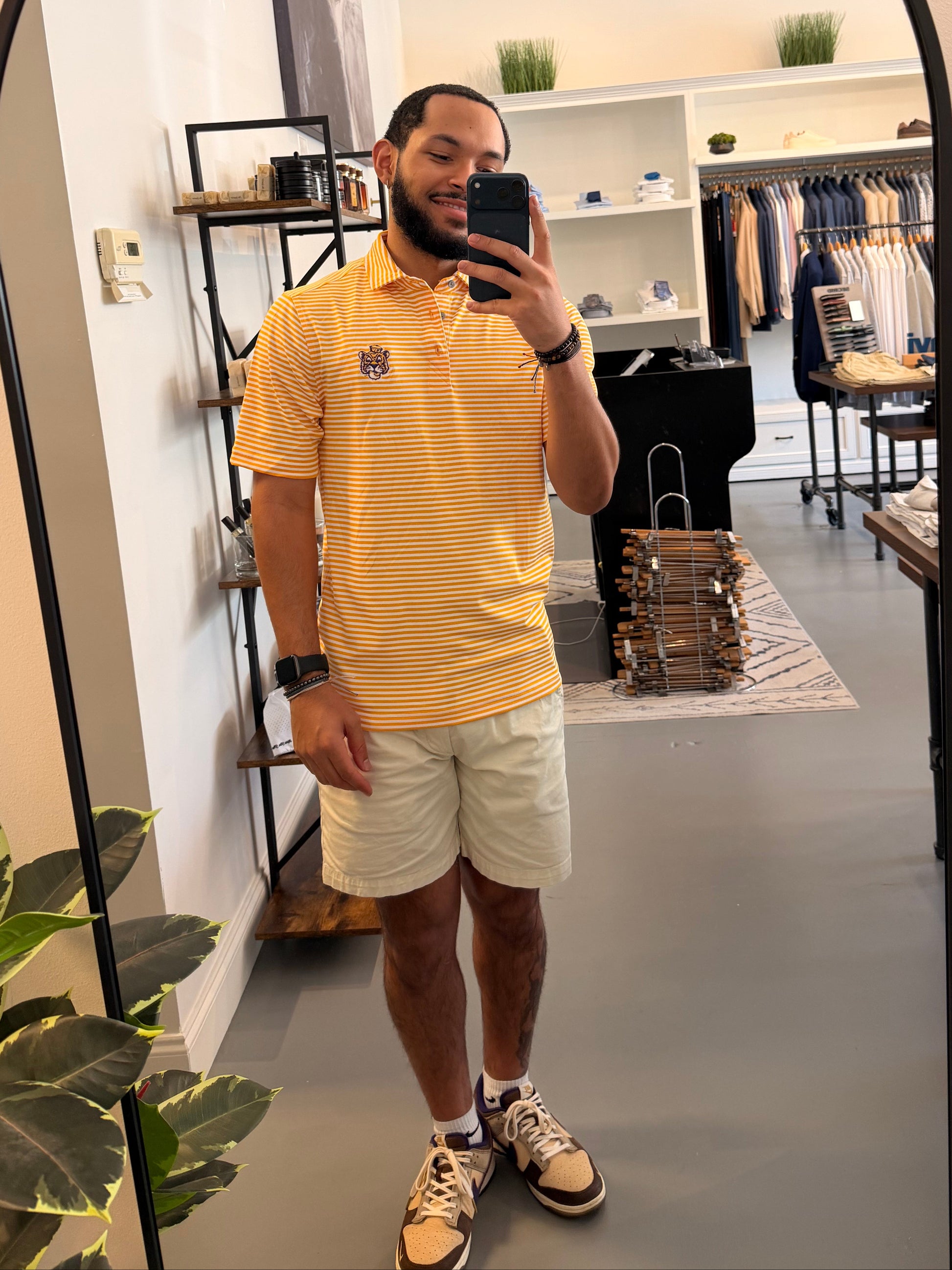 A man takes a mirror selfie in a clothing boutique wearing a LSU Sailor Mike the Tiger college gold and white striped polo shirt, beige shorts, and sneakers. The boutique features a large arched floor mirror with a black frame, white walls, and clothing racks in the background.