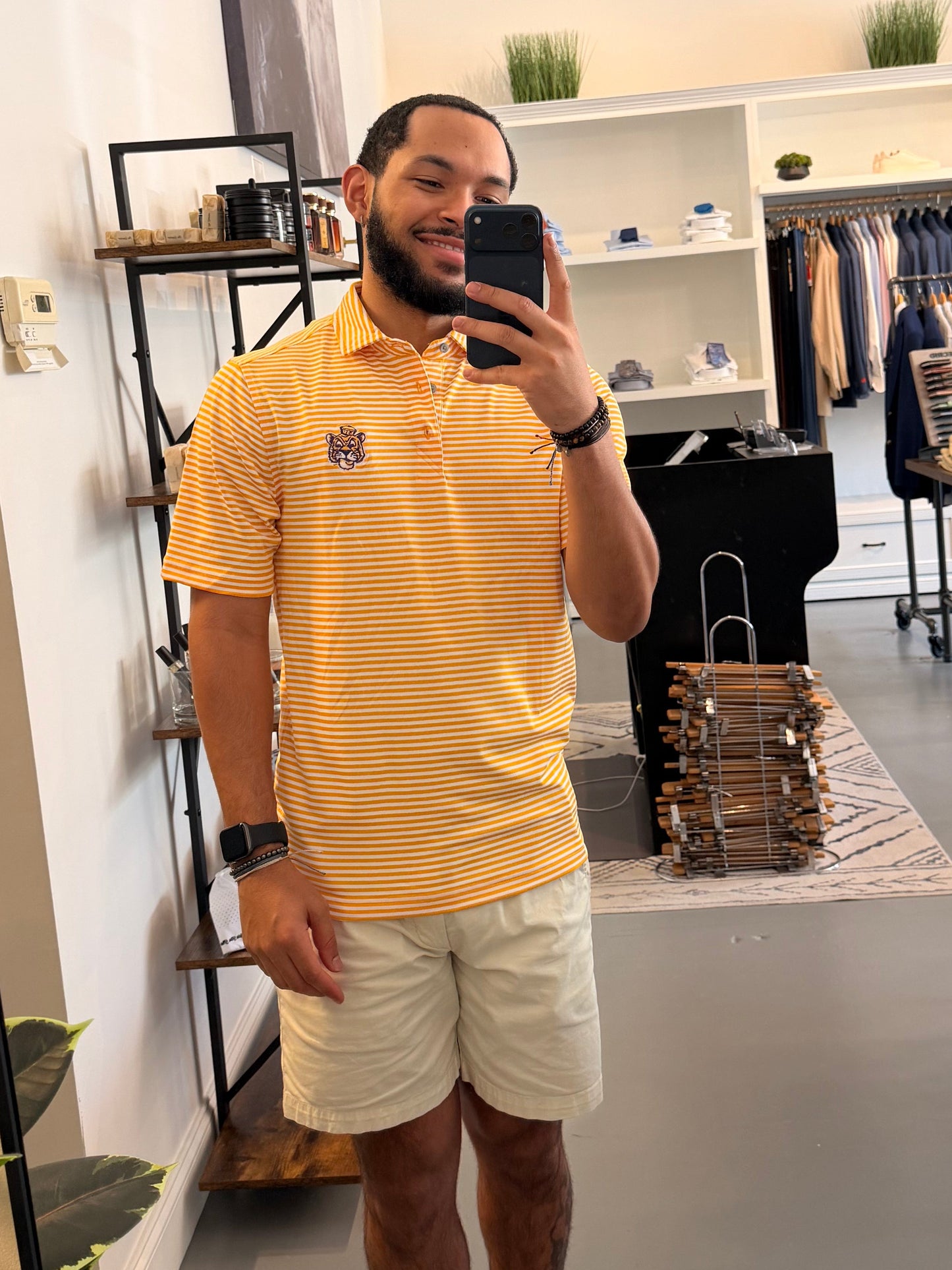 A man takes a mirror selfie in a clothing boutique wearing a LSU Sailor Mike the Tiger college gold and white striped polo shirt, beige shorts, and sneakers. The boutique features a large arched floor mirror with a black frame, white walls, and clothing racks in the background.