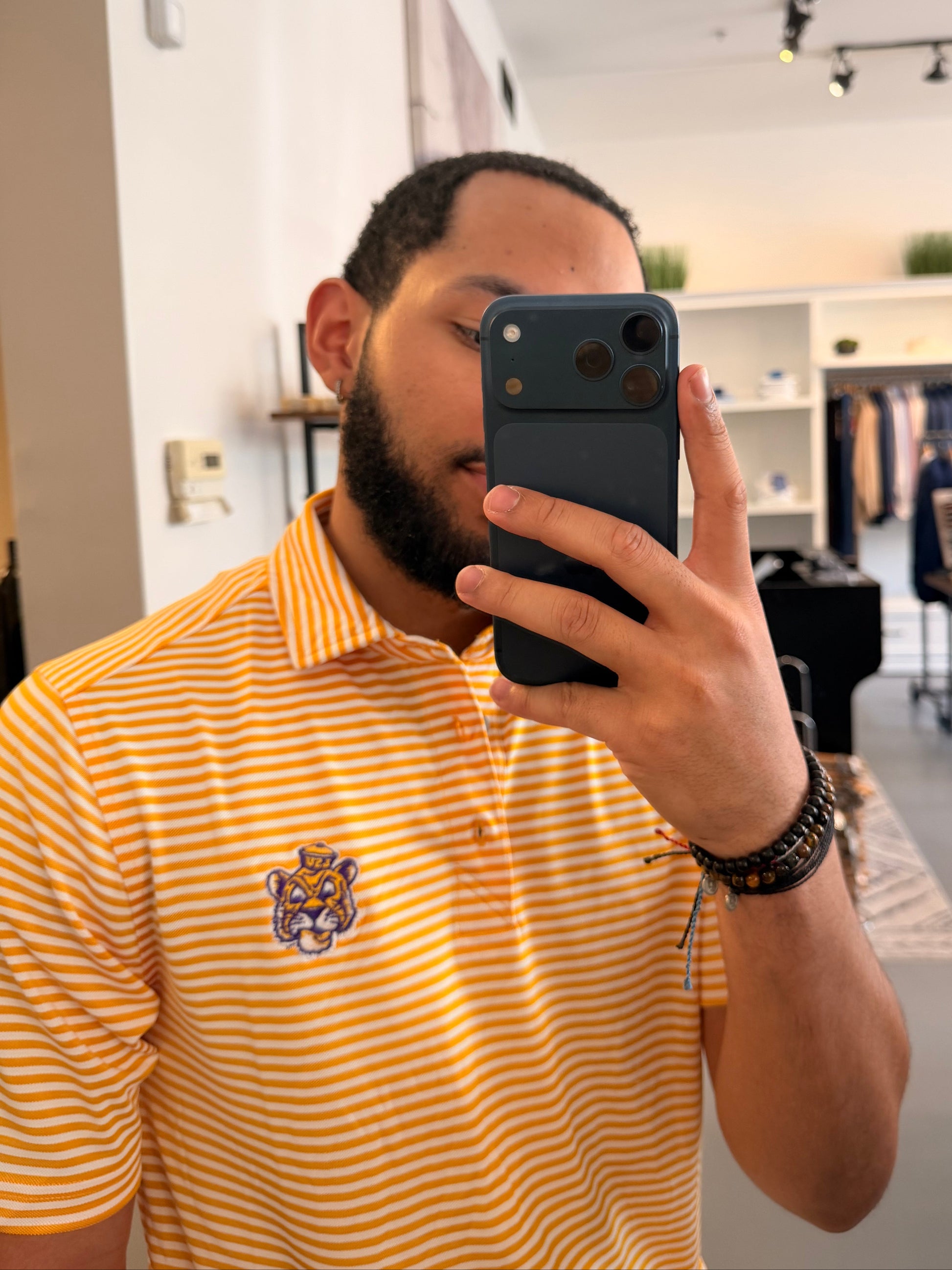 A man takes a mirror selfie in a clothing boutique wearing a LSU Sailor Mike the Tiger college gold and white striped polo shirt, beige shorts, and sneakers. The boutique features a large arched floor mirror with a black frame, white walls, and clothing racks in the background.