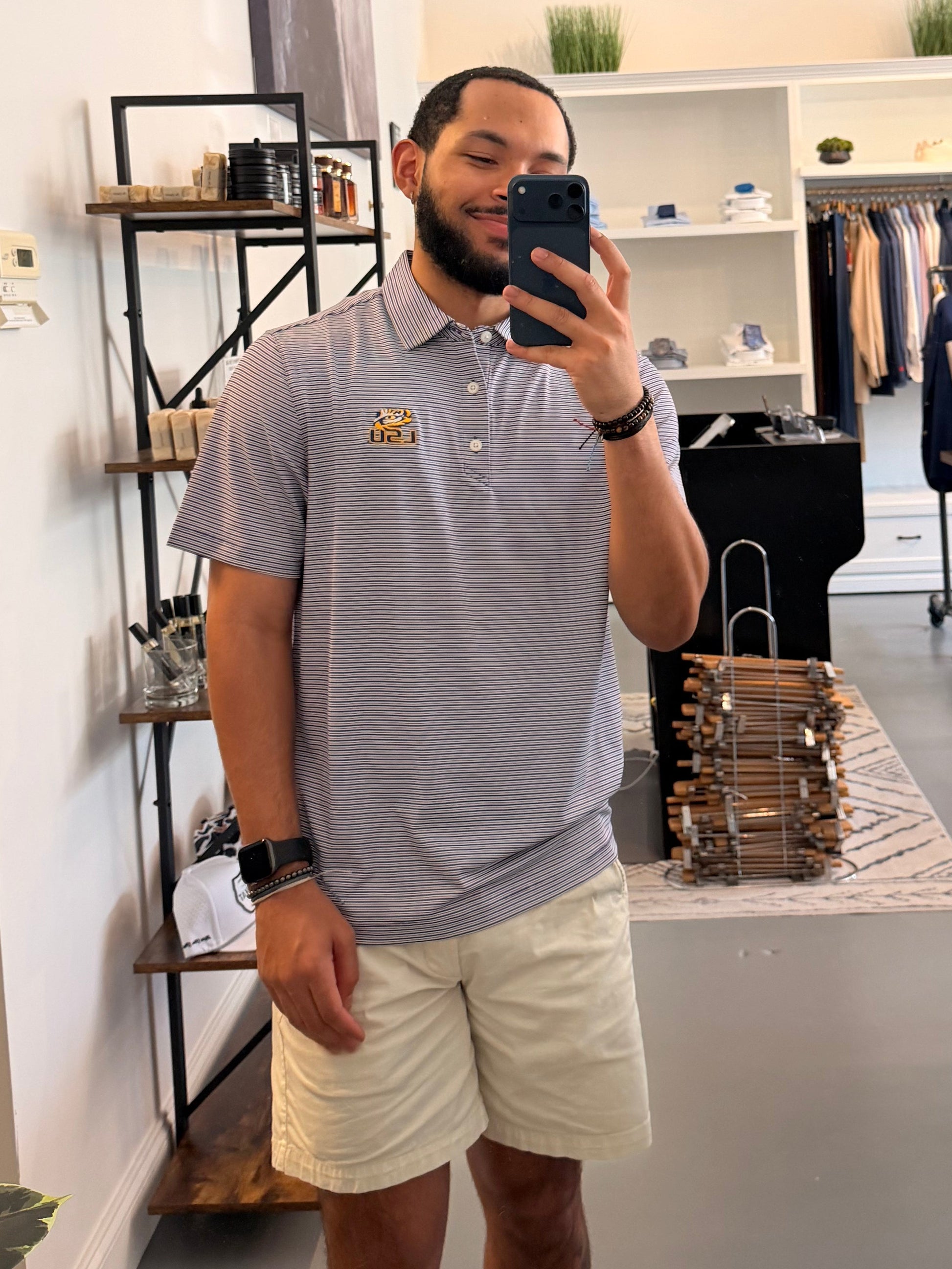 A man takes a mirror selfie in a boutique, wearing a College purple and white striped polo shirt with a LSU tiger EYE gold embroidered logo on the chest, paired with beige shorts.
