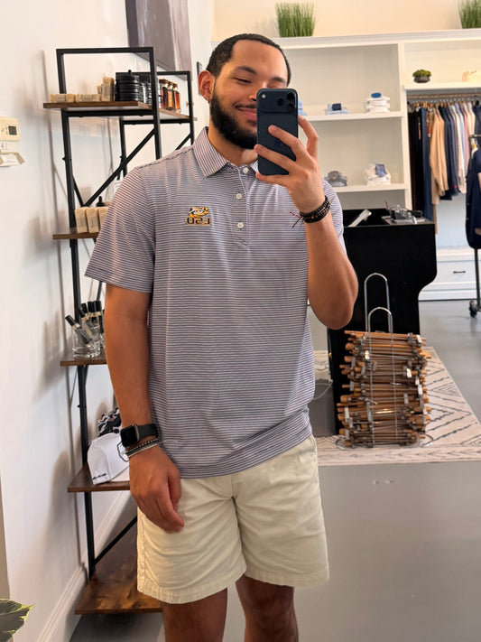 A man takes a mirror selfie in a boutique, wearing a College purple and white striped polo shirt with a LSU tiger EYE gold embroidered logo on the chest, paired with beige shorts.