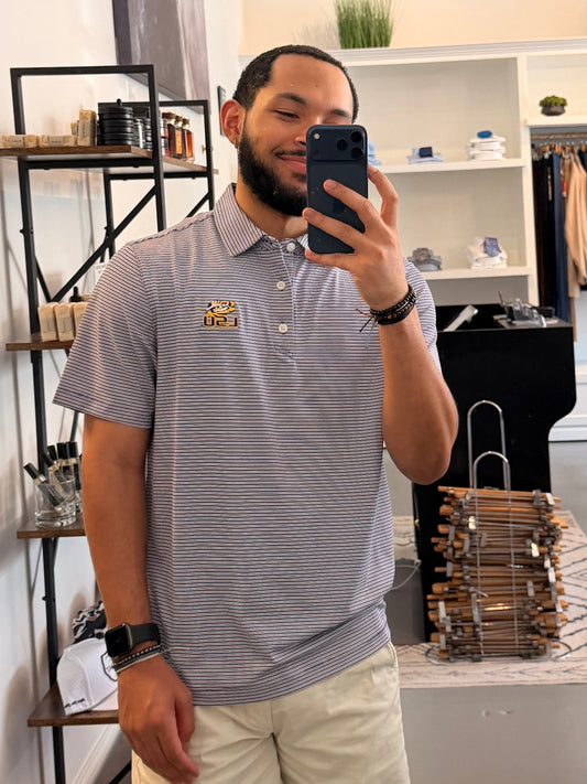 A man takes a mirror selfie in a boutique, wearing a College purple and white striped polo shirt with a LSU tiger EYE gold embroidered logo on the chest, paired with beige shorts.