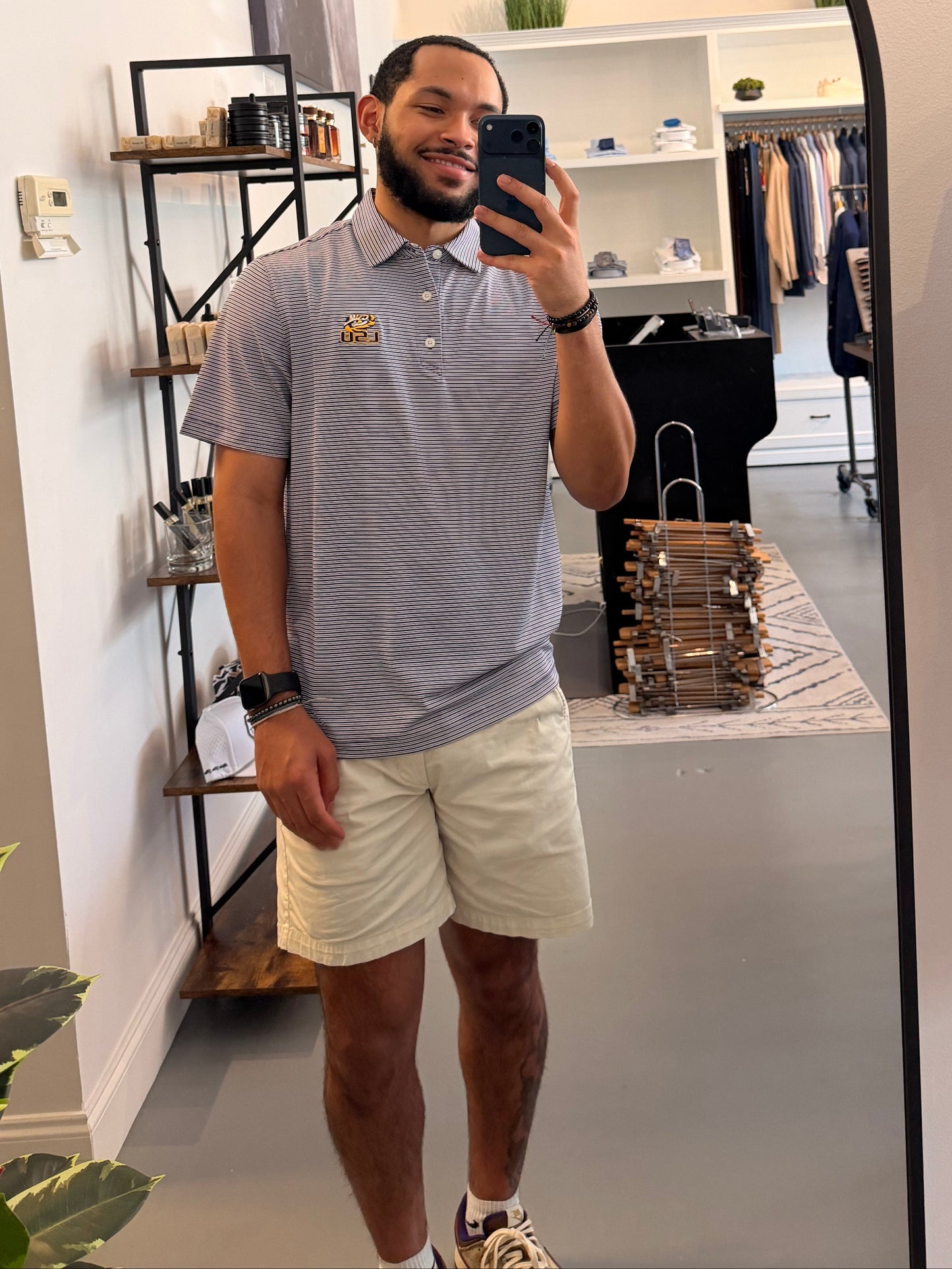 A man takes a mirror selfie in a boutique, wearing a College purple and white striped polo shirt with a LSU tiger EYE gold embroidered logo on the chest, paired with beige shorts.