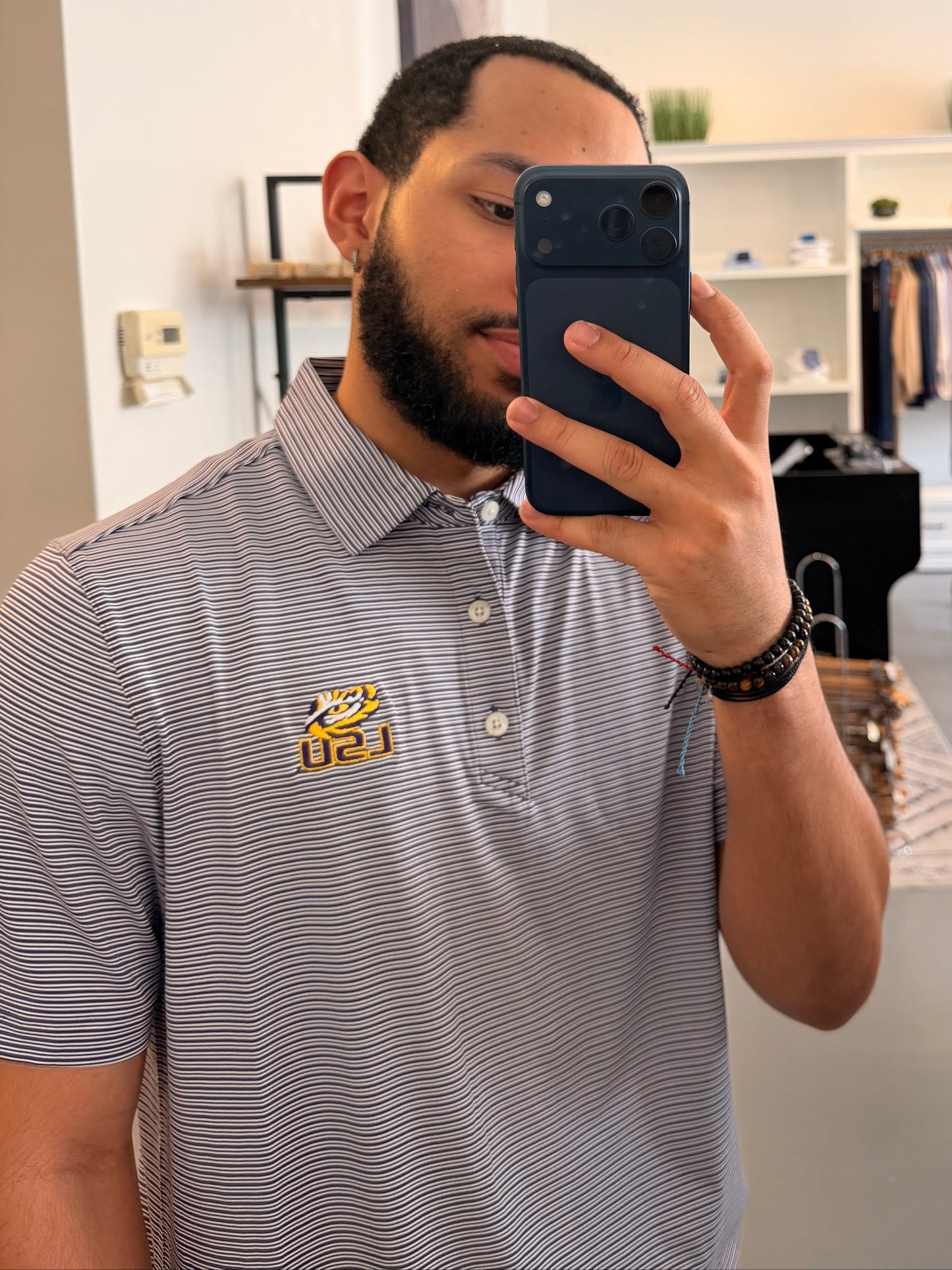 A man takes a mirror selfie in a boutique, wearing a College purple and white striped polo shirt with a LSU tiger EYE gold embroidered logo on the chest, paired with beige shorts.