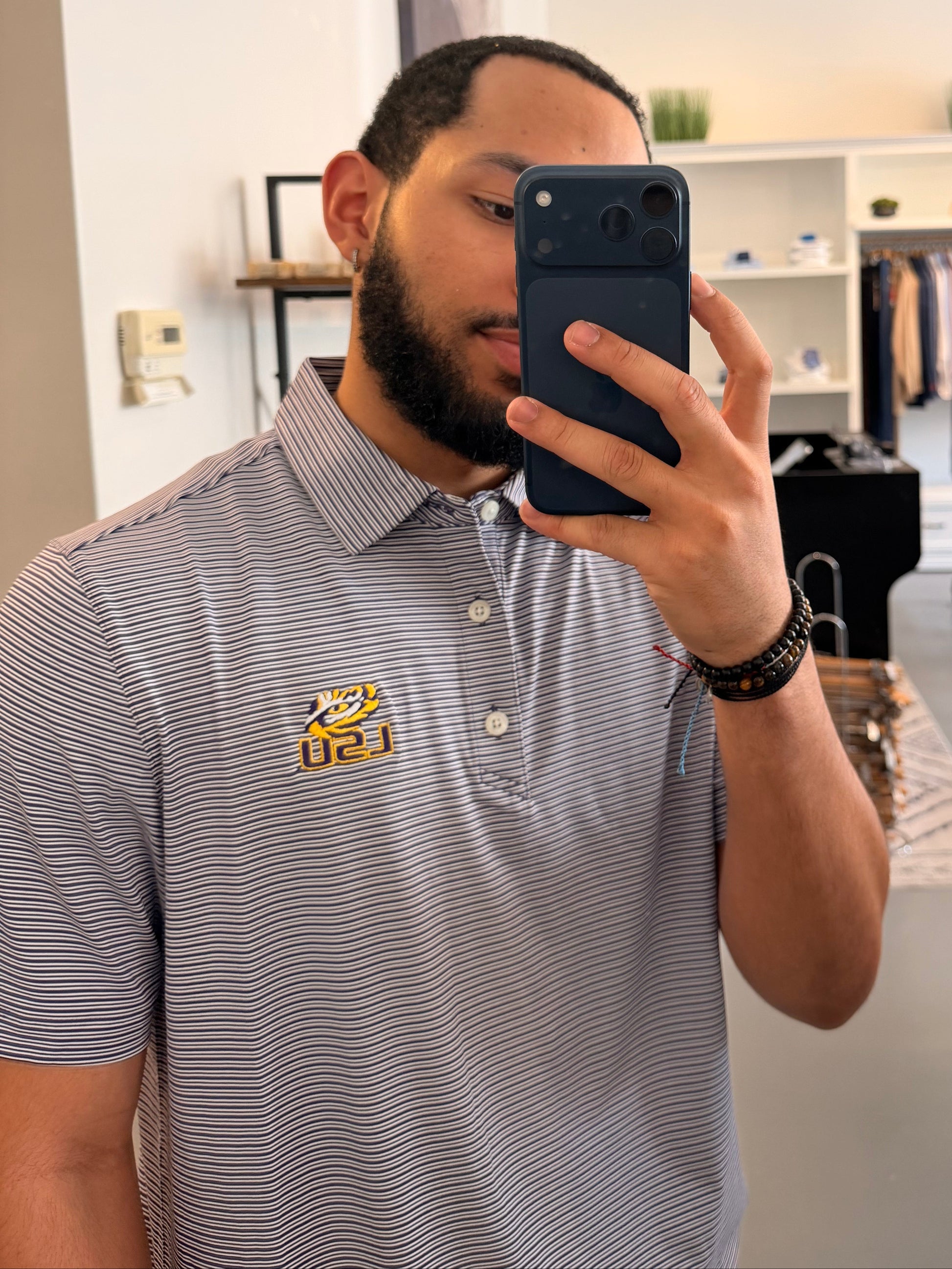 A man takes a mirror selfie in a boutique, wearing a College purple and white striped polo shirt with a LSU tiger EYE gold embroidered logo on the chest, paired with beige shorts.
