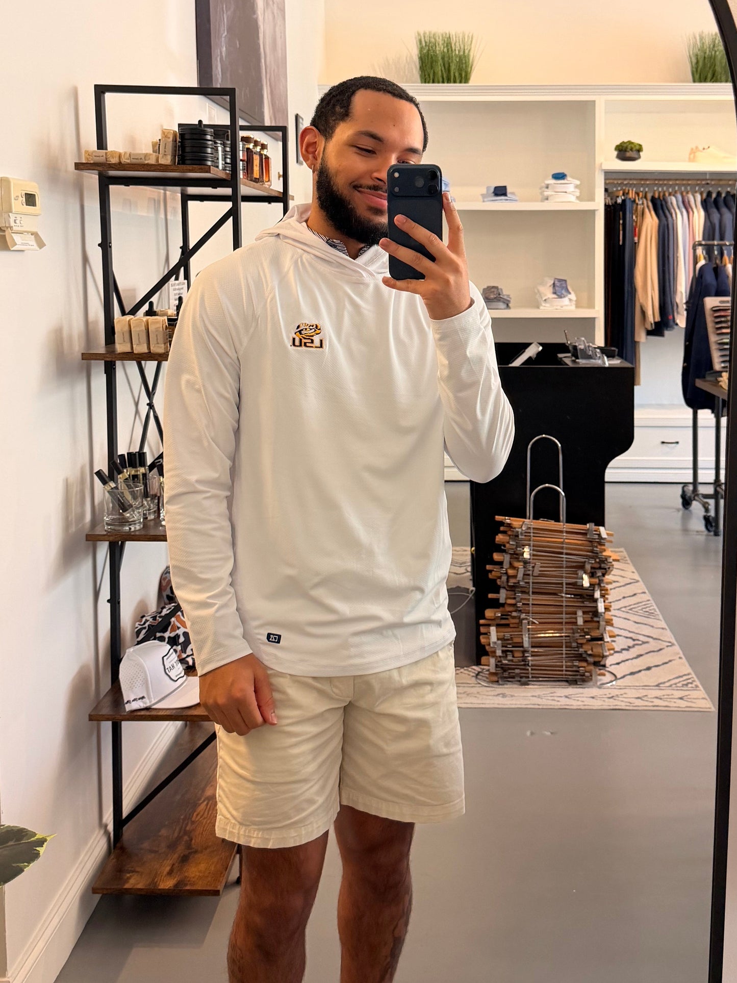 A man takes a mirror selfie in a boutique wearing a TIger Eye LSU logo white long-sleeve hoodie with a small LSU Tiger gold logo on the chest, beige shorts, and beige-and-black sneakers. The boutique interior is visible behind him, including clothing racks, shelves, and a plant.