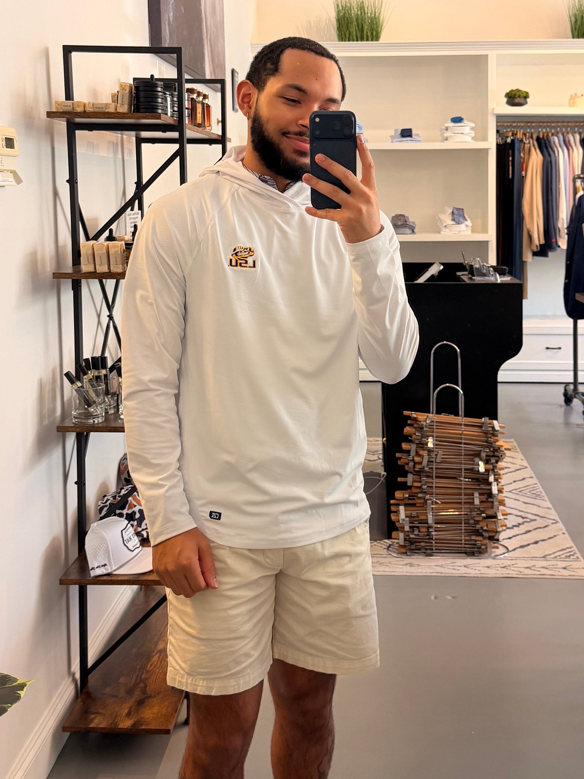 A man takes a mirror selfie in a boutique wearing a TIger Eye LSU logo white long-sleeve hoodie with a small LSU Tiger gold logo on the chest, beige shorts, and beige-and-black sneakers. The boutique interior is visible behind him, including clothing racks, shelves, and a plant.
