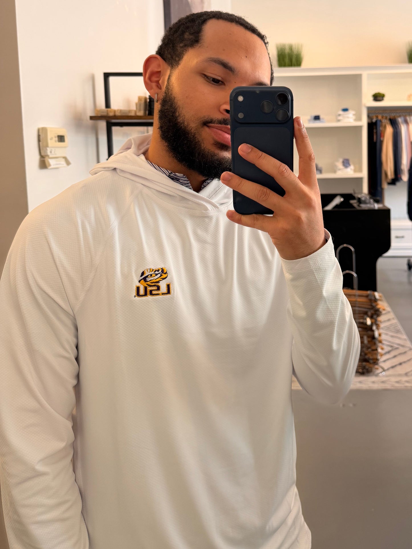 A man takes a mirror selfie in a boutique wearing a TIger Eye LSU logo white long-sleeve hoodie with a small LSU Tiger gold logo on the chest, beige shorts, and beige-and-black sneakers. The boutique interior is visible behind him, including clothing racks, shelves, and a plant.