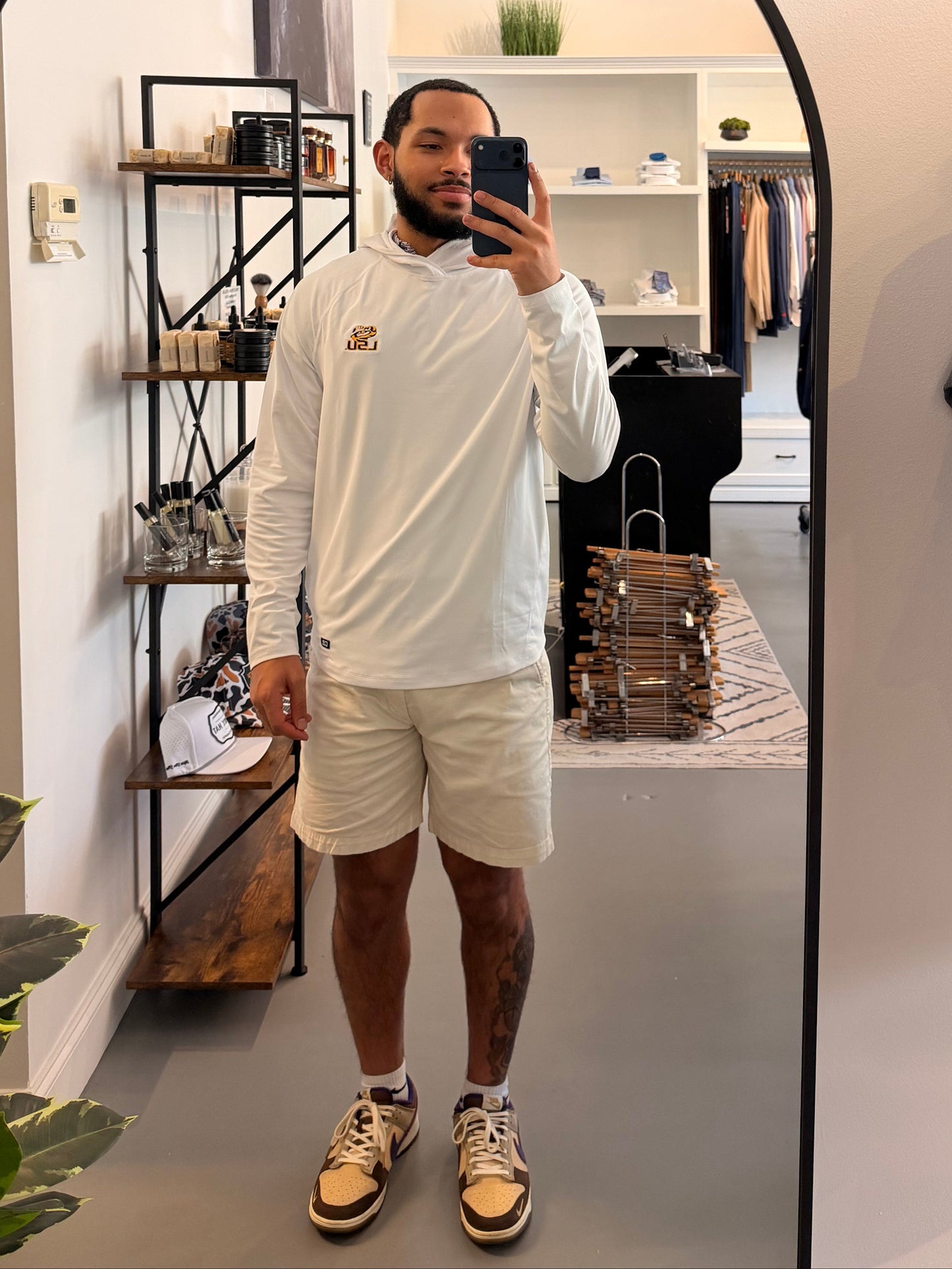 A man takes a mirror selfie in a boutique wearing a TIger Eye LSU logo white long-sleeve hoodie with a small LSU Tiger gold logo on the chest, beige shorts, and beige-and-black sneakers. The boutique interior is visible behind him, including clothing racks, shelves, and a plant.
