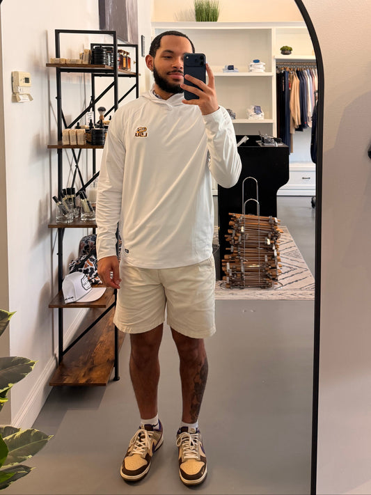 A man takes a mirror selfie in a boutique wearing a TIger Eye LSU logo white long-sleeve hoodie with a small LSU Tiger gold logo on the chest, beige shorts, and beige-and-black sneakers. The boutique interior is visible behind him, including clothing racks, shelves, and a plant.