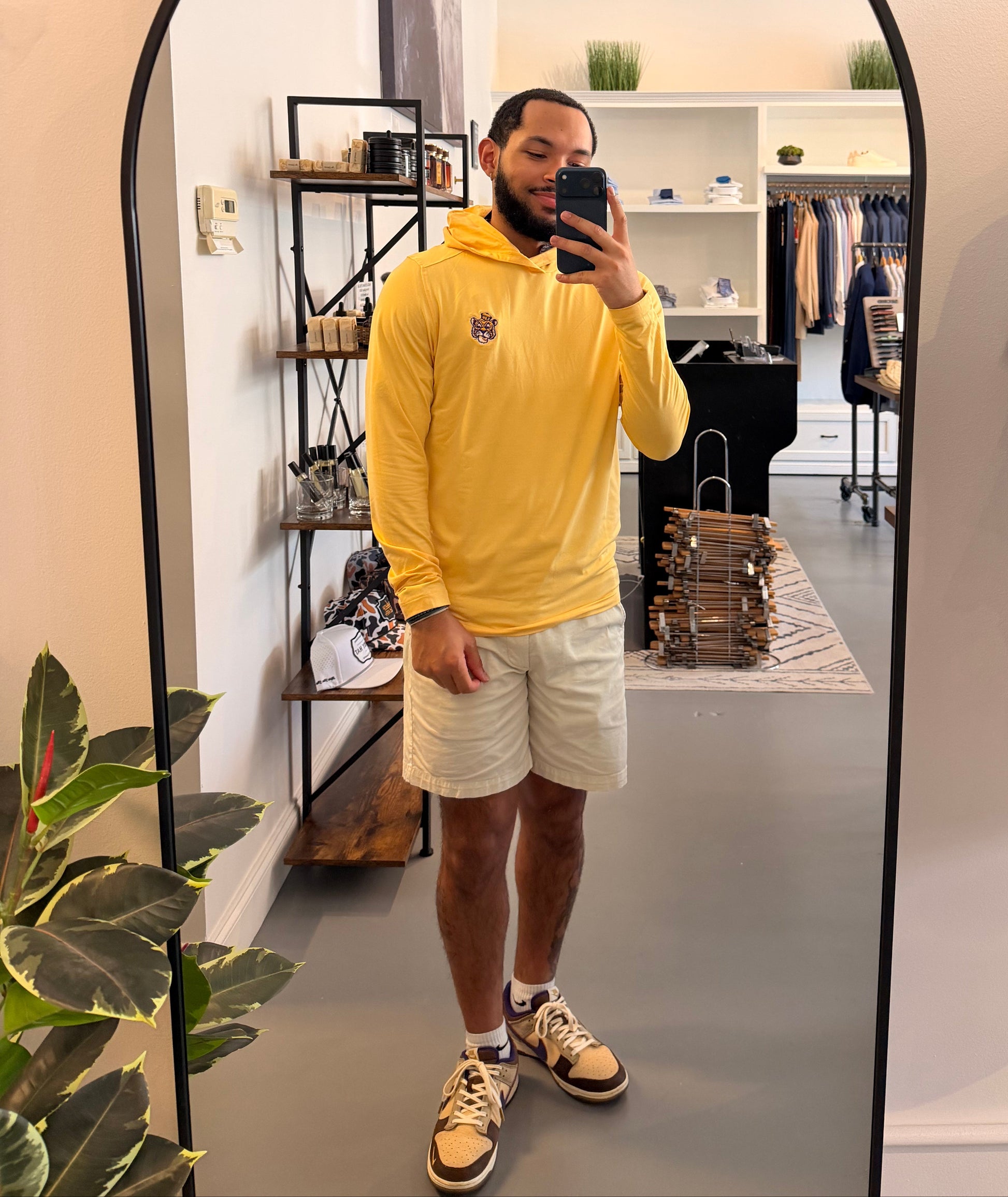 A man takes a mirror selfie in a boutique wearing a LSU Tiger college gold hoodie, beige shorts, and beige-and-brown sneakers. The boutique interior features a black-framed mirror, shelving, and clothing racks.