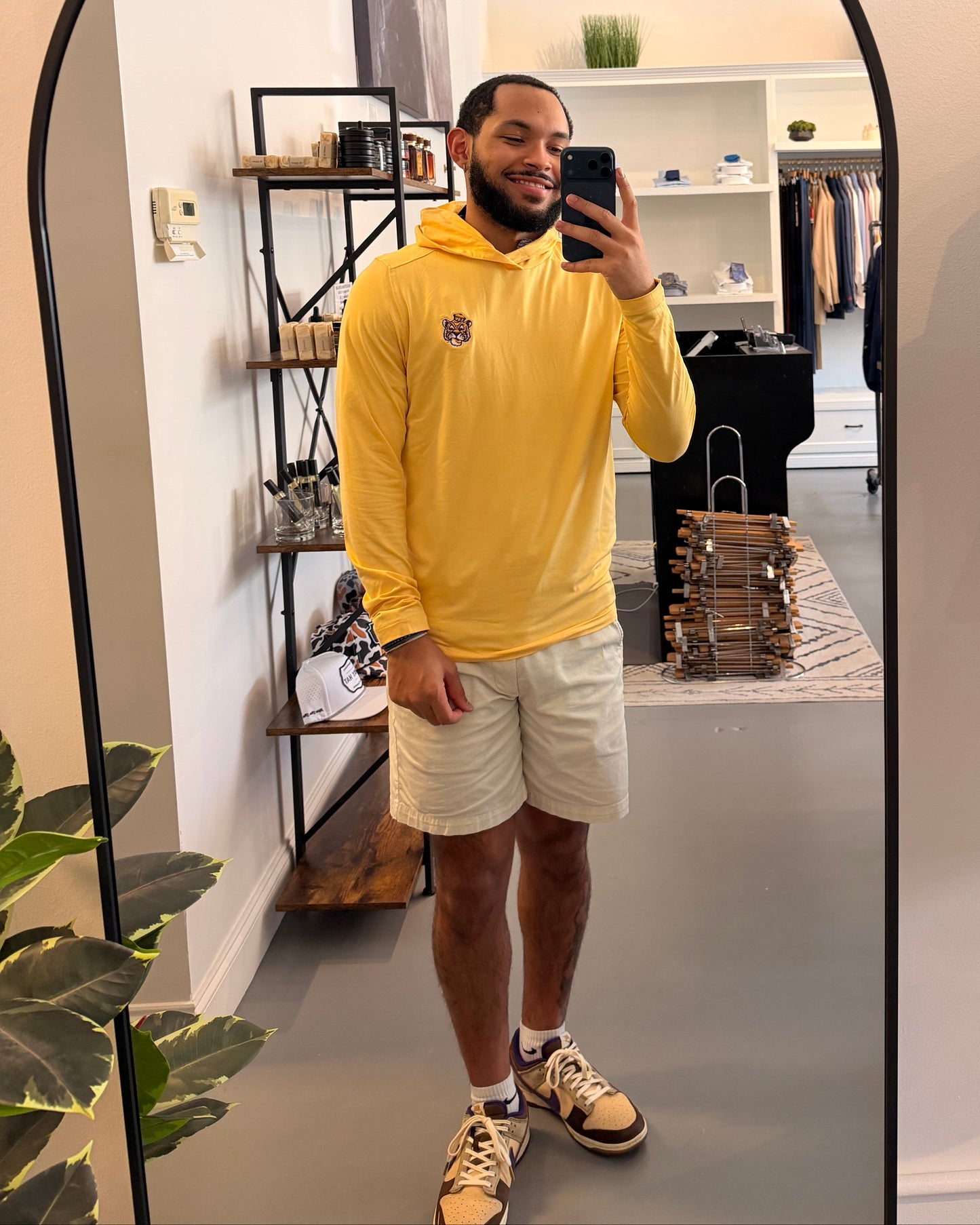 A man takes a mirror selfie in a boutique wearing a LSU Tigers college gold long-sleeve hoodie with a small LSU embroidered on the left chest, paired with beige shorts and beige-and-black sneakers.
