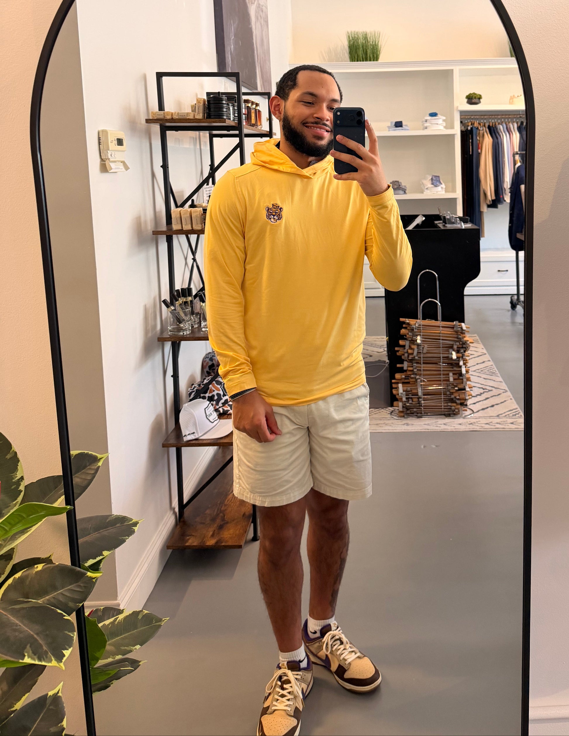 A man takes a mirror selfie in a boutique wearing a LSU Tigers college gold long-sleeve hoodie with a small LSU embroidered on the left chest, paired with beige shorts and beige-and-black sneakers.