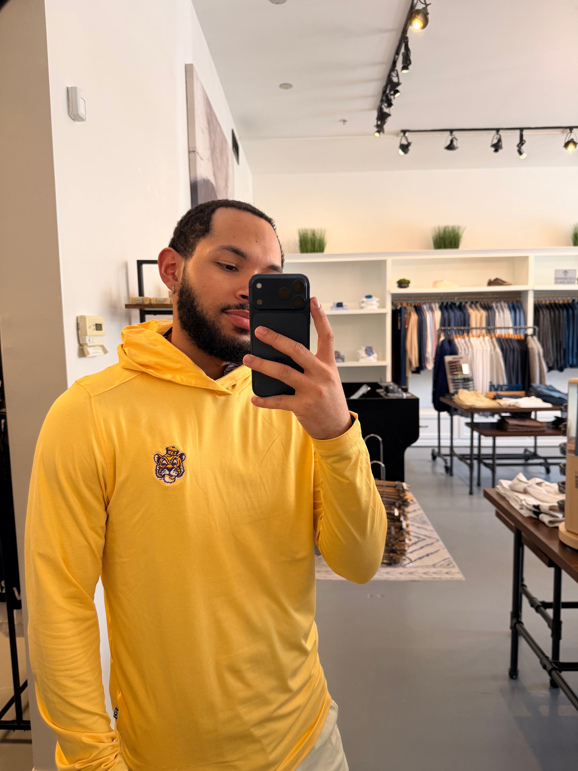 A man takes a mirror selfie in a boutique wearing a LSU Tigers college gold long-sleeve hoodie with a small LSU embroidered on the left chest, paired with beige shorts and beige-and-black sneakers.