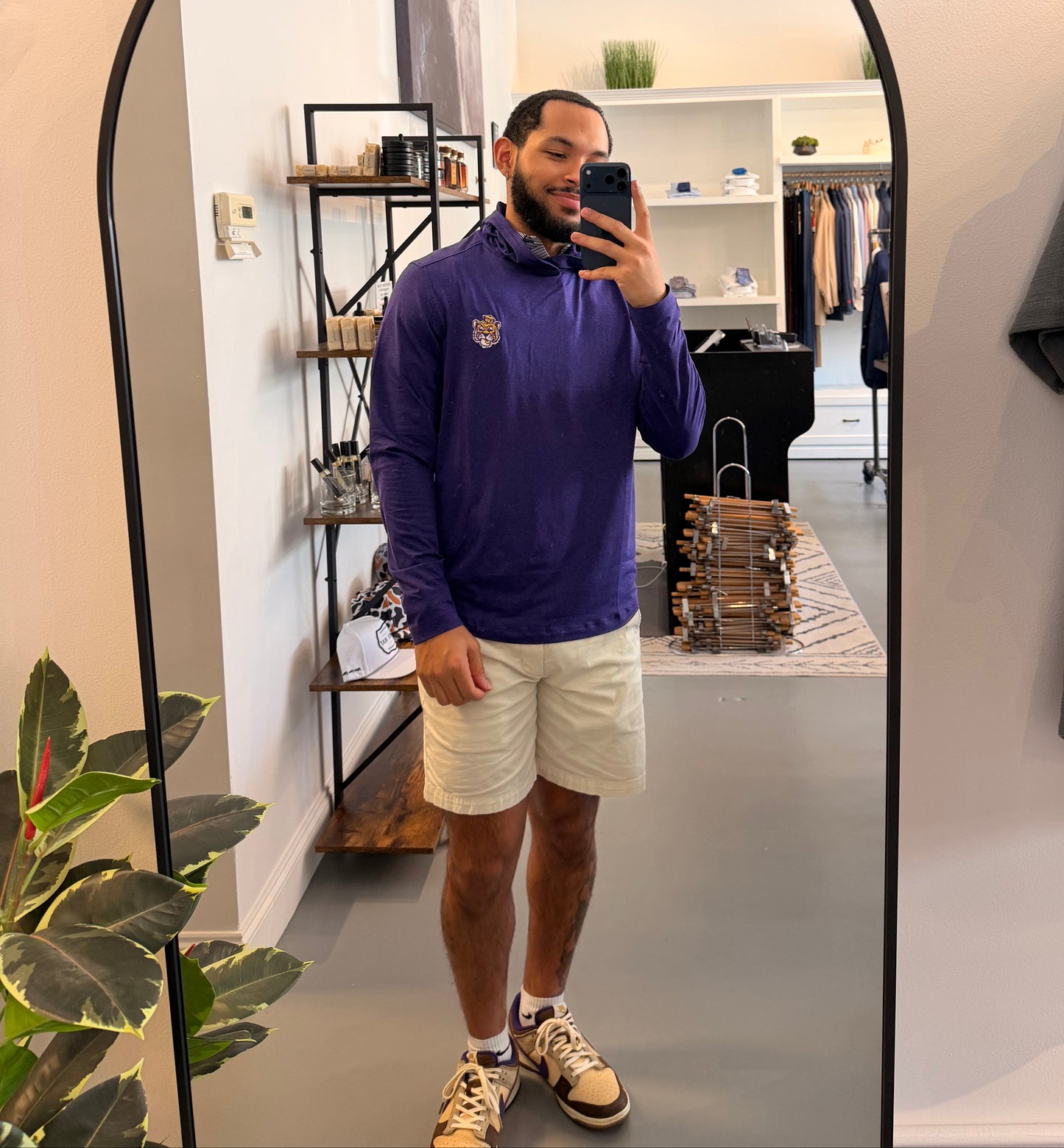 A man takes a mirror selfie in a boutique, wearing a LSU Tiger purple long-sleeve athletic pullover with a small gold crest on the left chest, paired with off-white shorts and sneakers. The boutique interior is visible in the background behind an arched floor mirror.