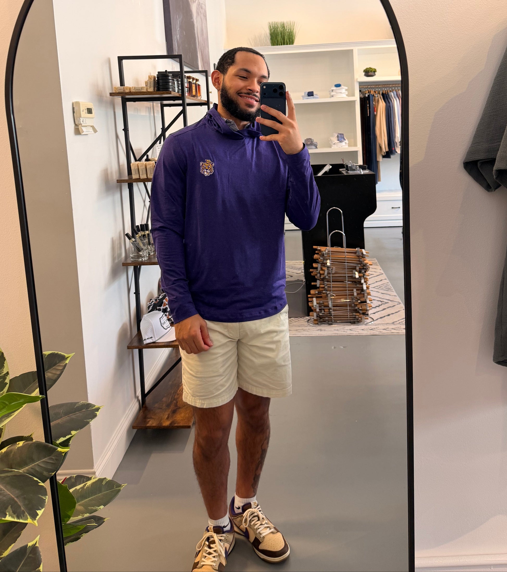 A man takes a mirror selfie in a boutique showing a LSU Tiger purple long-sleeve crewneck sweater, cream shorts, and sneakers. The boutique has a white arched floor mirror, shelves, and clothing racks.