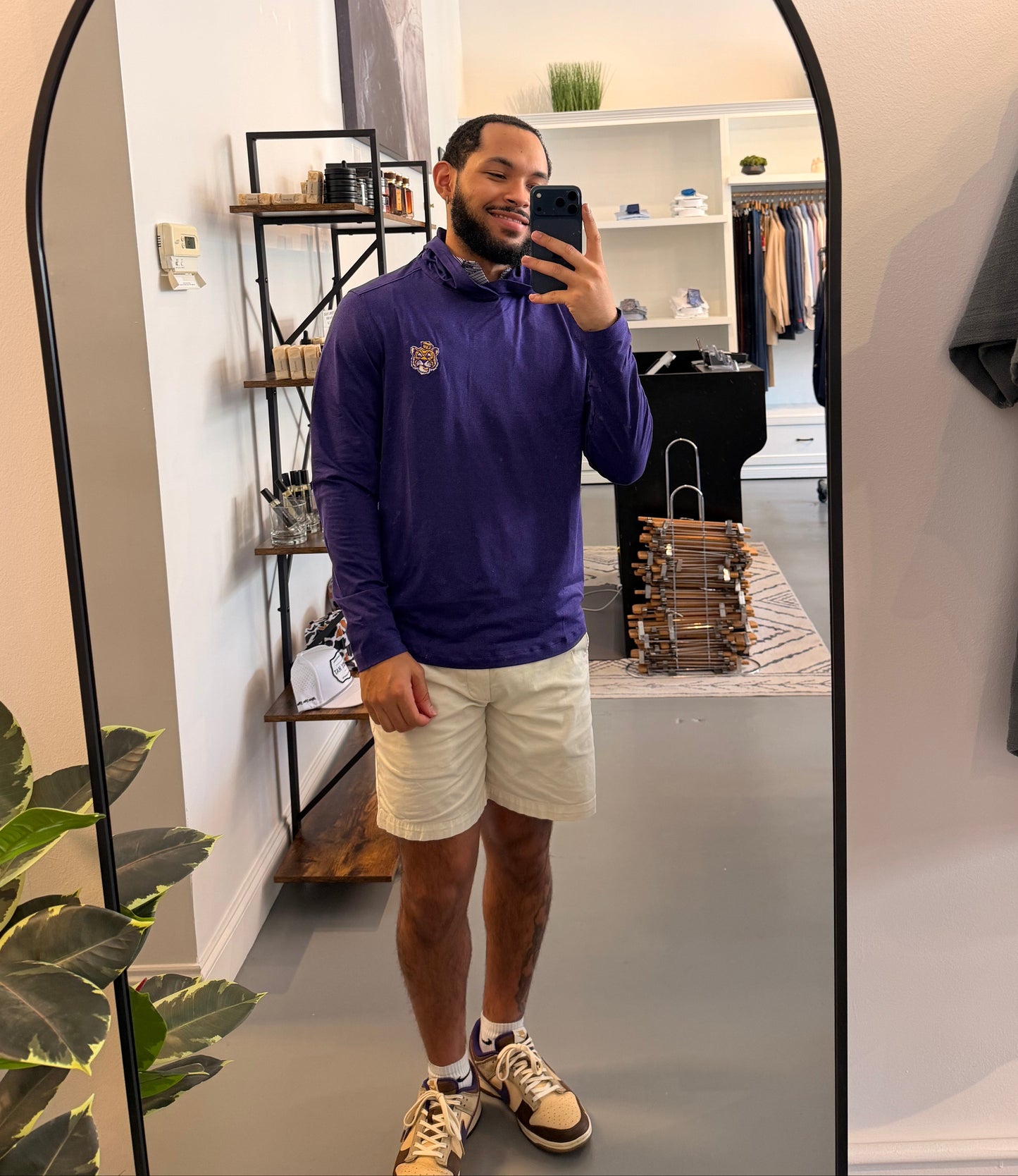 A man takes a mirror selfie in a boutique, wearing a LSU Tiger purple long-sleeve quarter-zip pullover, beige shorts, and sneakers. The boutique interior is visible in the background behind the large arched mirror.