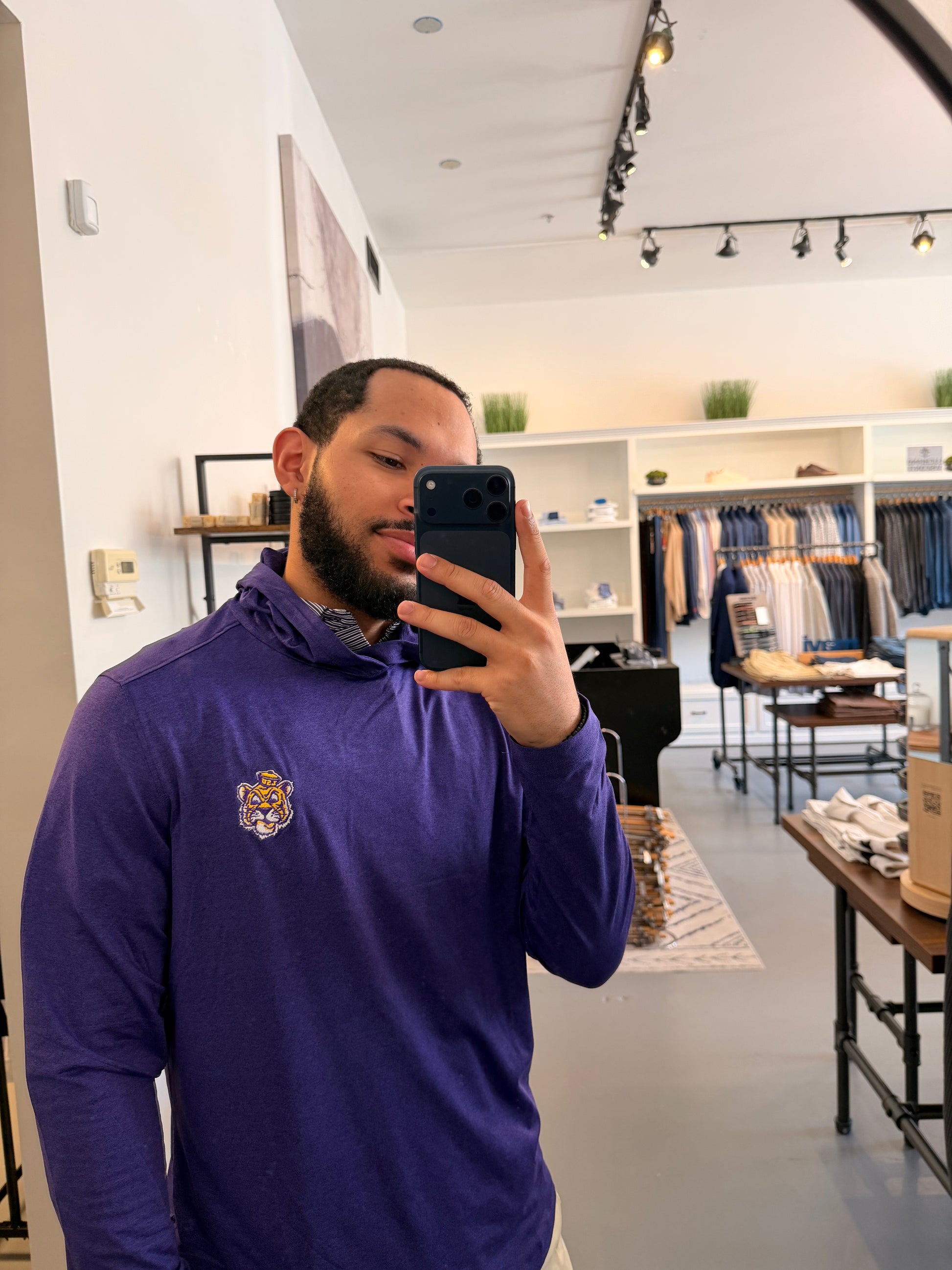 A man takes a mirror selfie in a clothing store, wearing a LSU Tiger purple long-sleeve quarter-zip pullover with a small embroidered crest on the left chest.