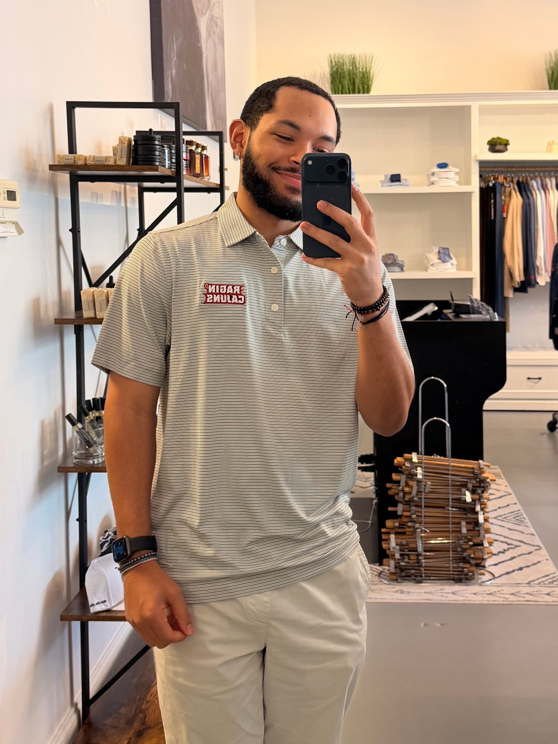 A man takes a mirror selfie in a clothing boutique, wearing a Louisiana ULL Ragin Cajuns light gray and white striped polo shirt with a Ragin Cajuns red logo on the chest and light beige pants. The photo is taken in front of a large arched black-framed mirror, with store fixtures and clothing racks visible in the background.