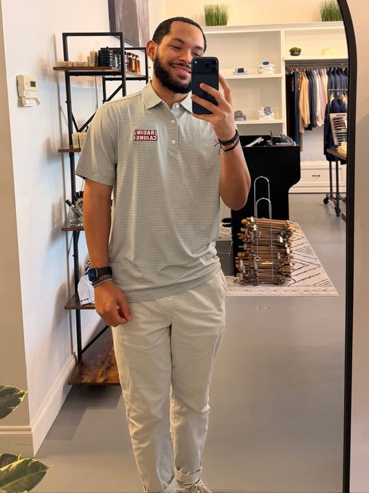 A man takes a mirror selfie in a clothing boutique, wearing a Louisiana ULL Ragin Cajuns light gray and white striped polo shirt with a Ragin Cajuns red logo on the chest and light beige pants. The photo is taken in front of a large arched black-framed mirror, with store fixtures and clothing racks visible in the background.