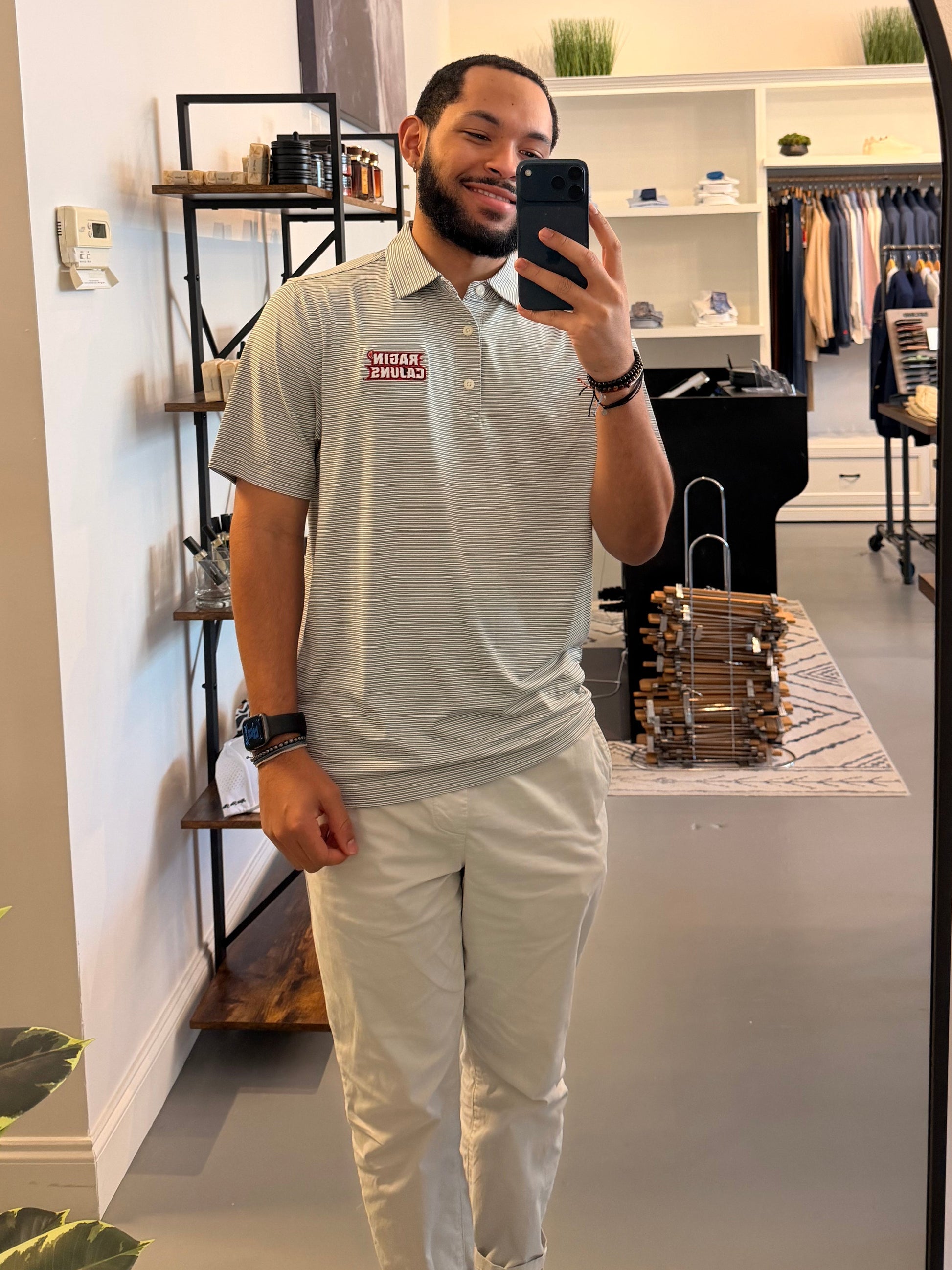 A man takes a mirror selfie in a clothing boutique, wearing a Louisiana ULL Ragin Cajuns light gray and white striped polo shirt with a Ragin Cajuns red logo on the chest and light beige pants. The photo is taken in front of a large arched black-framed mirror, with store fixtures and clothing racks visible in the background.