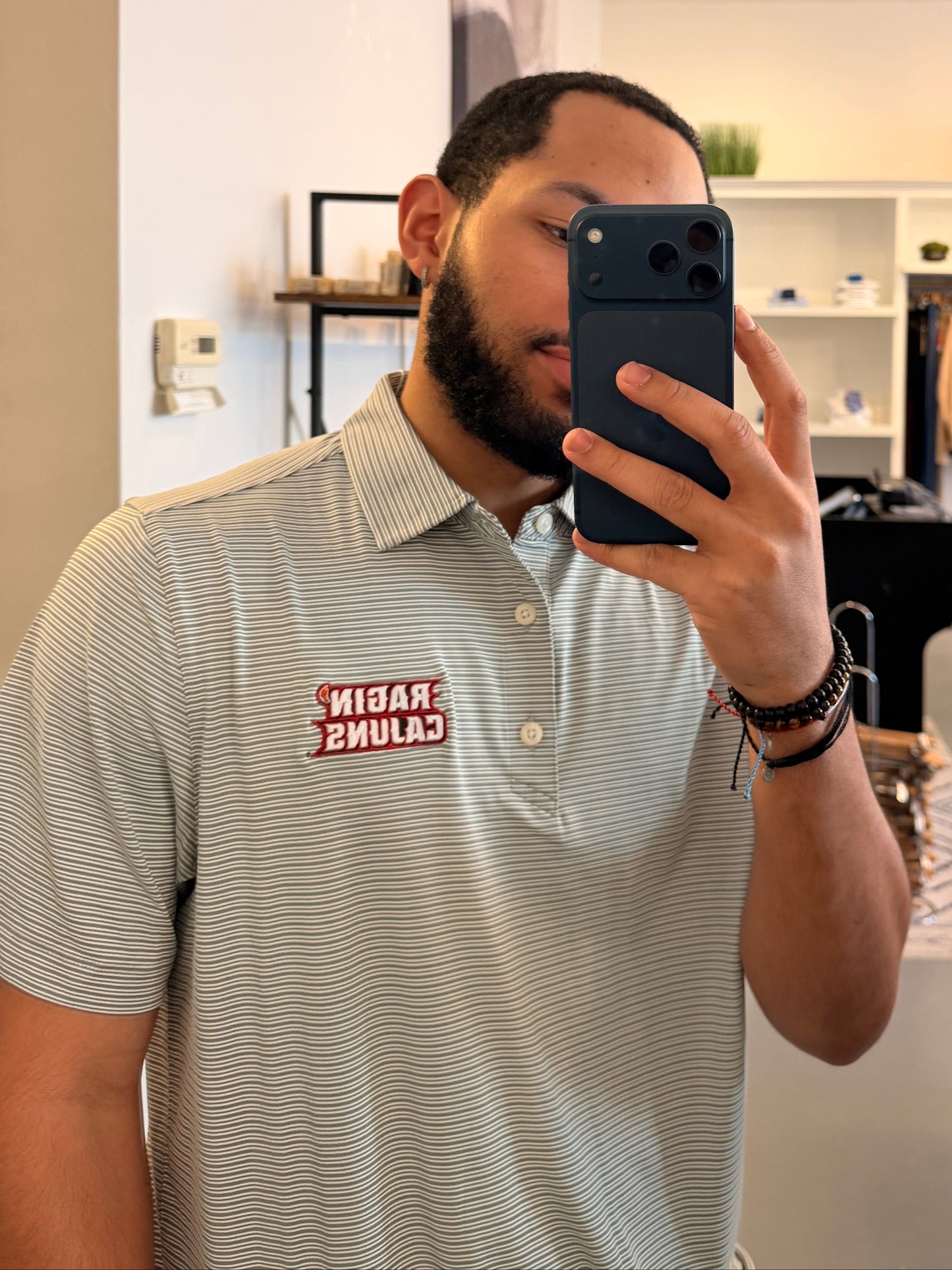 A man takes a mirror selfie in a clothing boutique, wearing a Louisiana ULL Ragin Cajuns light gray and white striped polo shirt with a Ragin Cajuns red logo on the chest and light beige pants. The photo is taken in front of a large arched black-framed mirror, with store fixtures and clothing racks visible in the background.