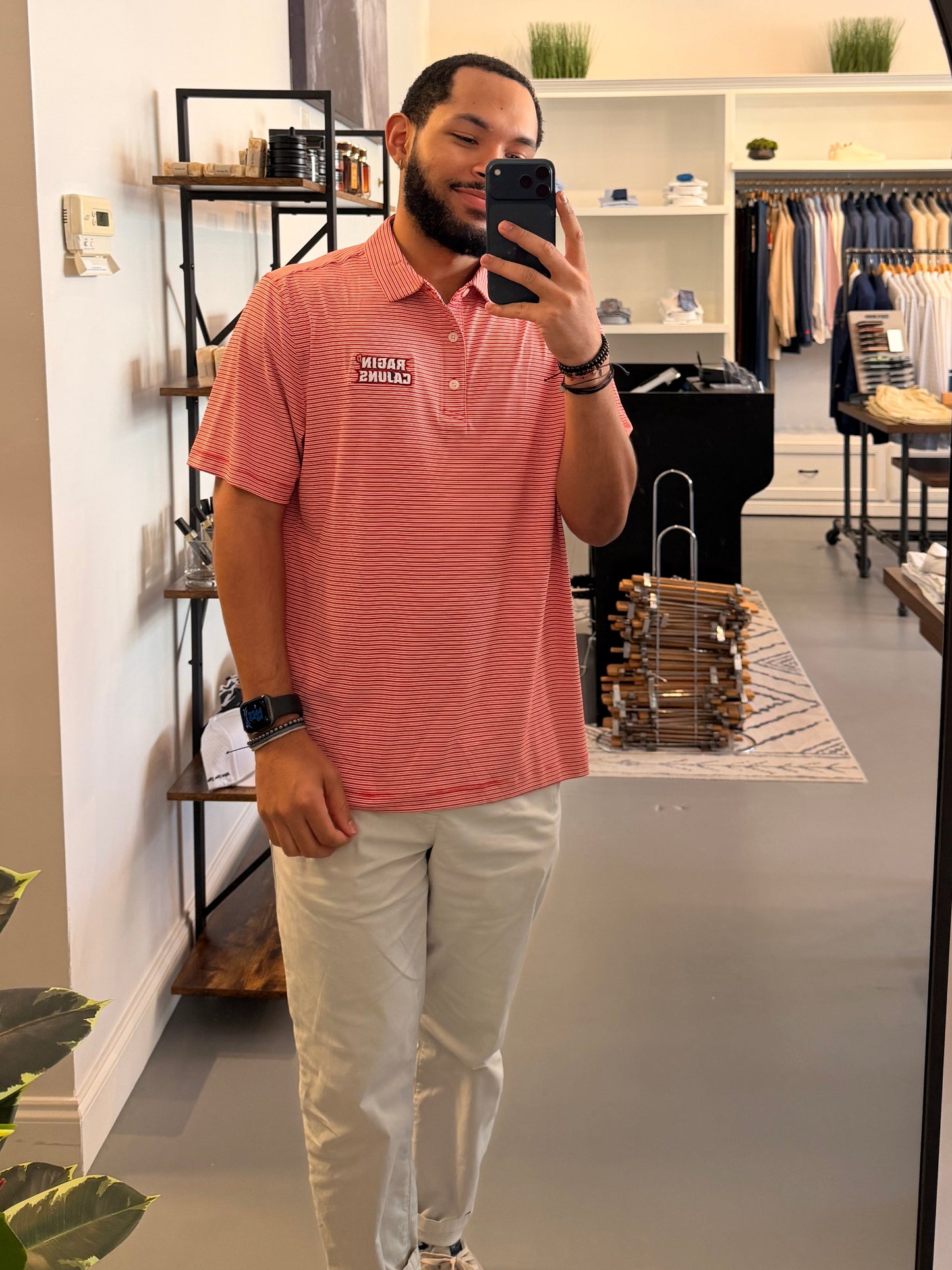 A man takes a mirror selfie in a clothing boutique wearing a University of Louisiana-Lafayette Ragin Cajuns red and white striped polo shirt with a Ragin Cajuns embroidered logo on the chest, paired with light beige pants.