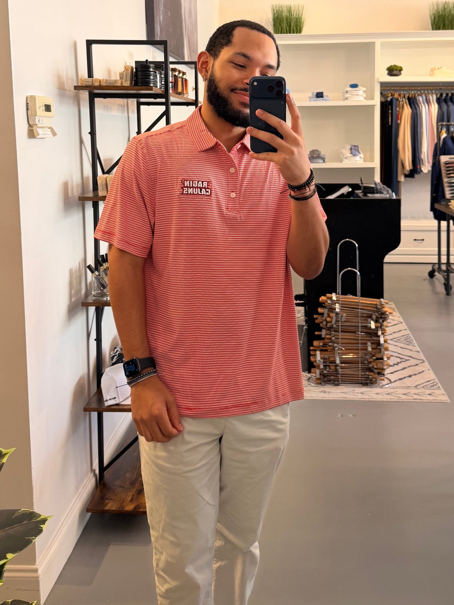 A man takes a mirror selfie in a clothing boutique wearing a University of Louisiana-Lafayette Ragin Cajuns red and white striped polo shirt with a Ragin Cajuns embroidered logo on the chest, paired with light beige pants.