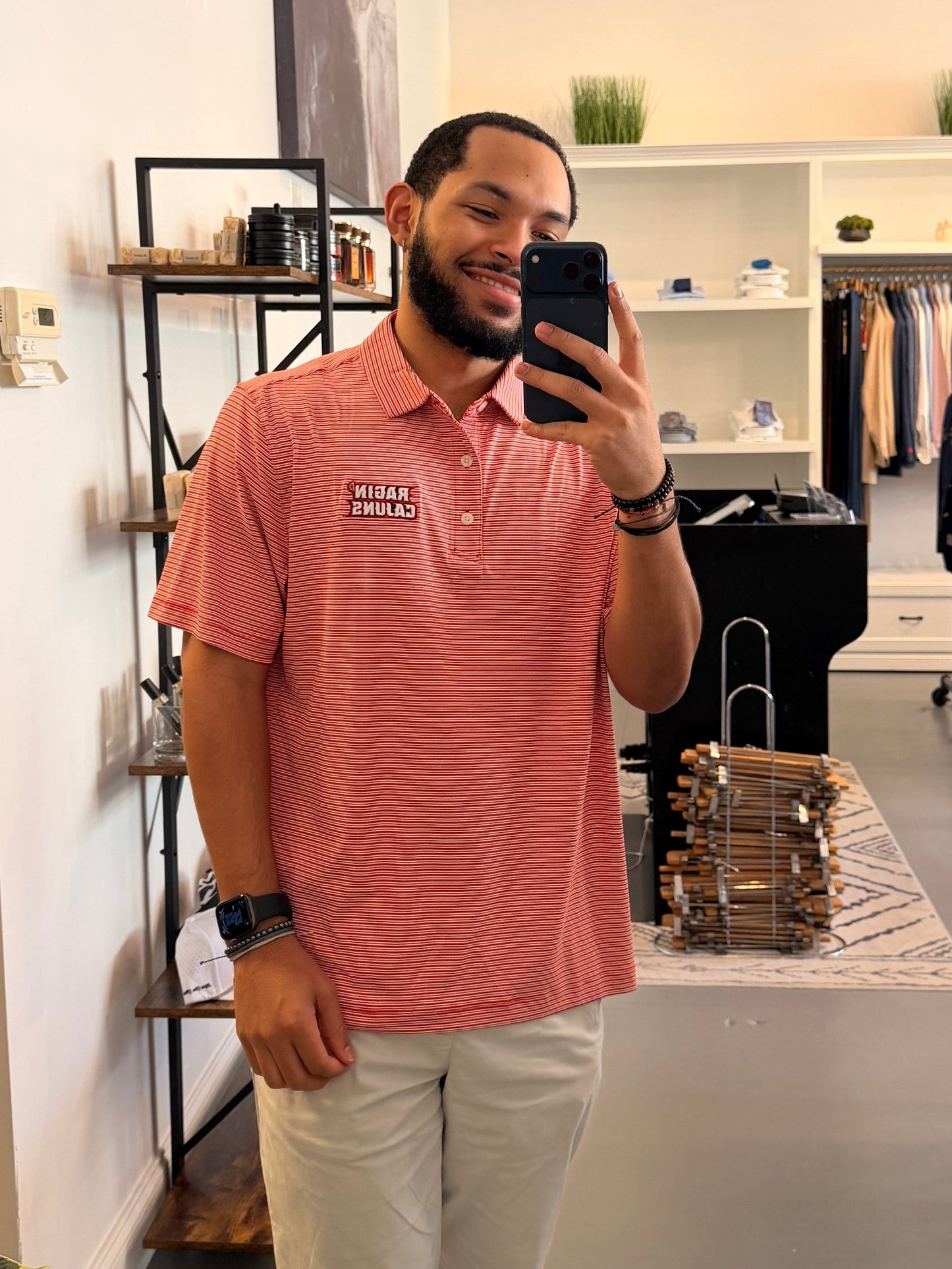 A man takes a mirror selfie in a clothing boutique wearing a University of Louisiana-Lafayette Ragin Cajuns red and white striped polo shirt with a Ragin Cajuns embroidered logo on the chest, paired with light beige pants.