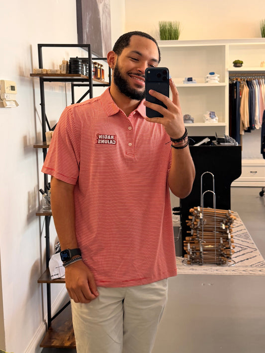 A man takes a mirror selfie in a clothing boutique wearing a University of Louisiana-Lafayette Ragin Cajuns red and white striped polo shirt with a Ragin Cajuns embroidered logo on the chest, paired with light beige pants.