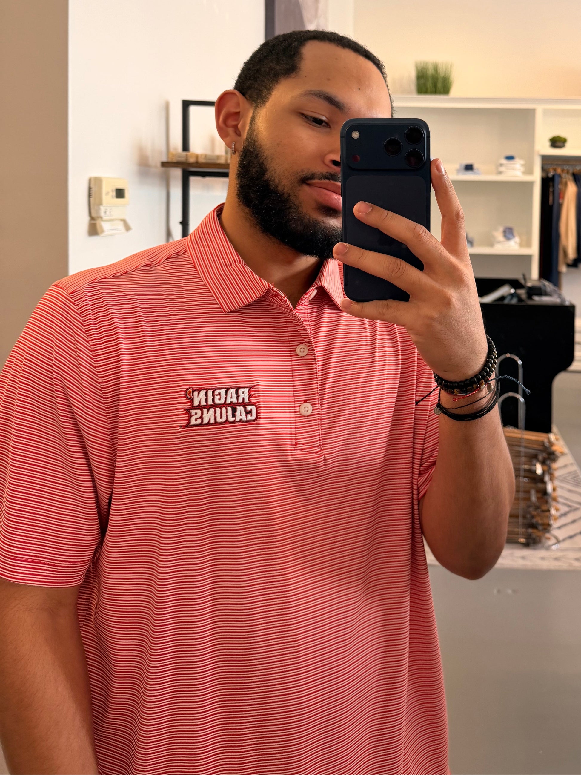 A man takes a mirror selfie in a clothing boutique wearing a University of Louisiana-Lafayette Ragin Cajuns red and white striped polo shirt with a Ragin Cajuns embroidered logo on the chest, paired with light beige pants.