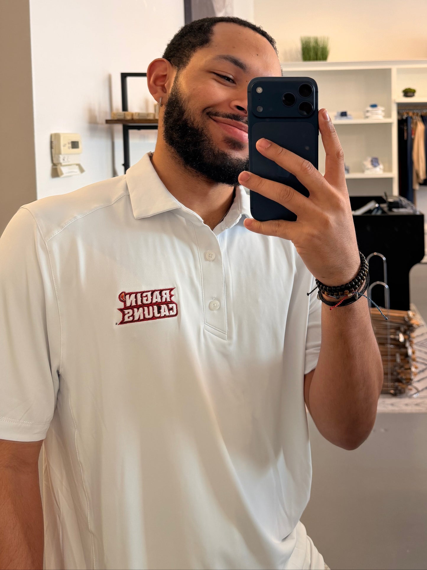 A man takes a mirror selfie in a clothing boutique, wearing a University of Louisiana-Lafayette Ragin Cajuns white polo shirt with a Ragin Cajuns red embroidered logo on the chest and white pants. The boutique has a modern interior with a large arched mirror, clothing racks, and shelves.