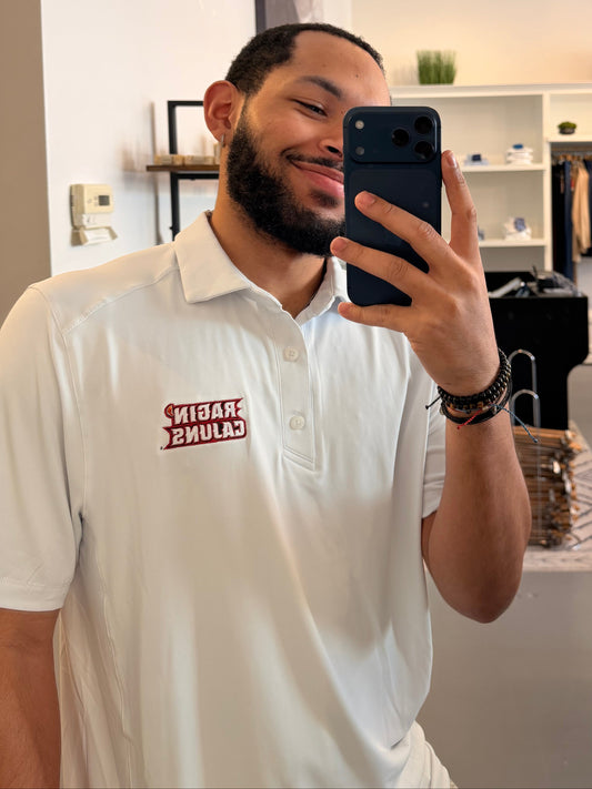A man takes a mirror selfie in a clothing boutique, wearing a University of Louisiana-Lafayette Ragin Cajuns white polo shirt with a Ragin Cajuns red embroidered logo on the chest and white pants. The boutique has a modern interior with a large arched mirror, clothing racks, and shelves.