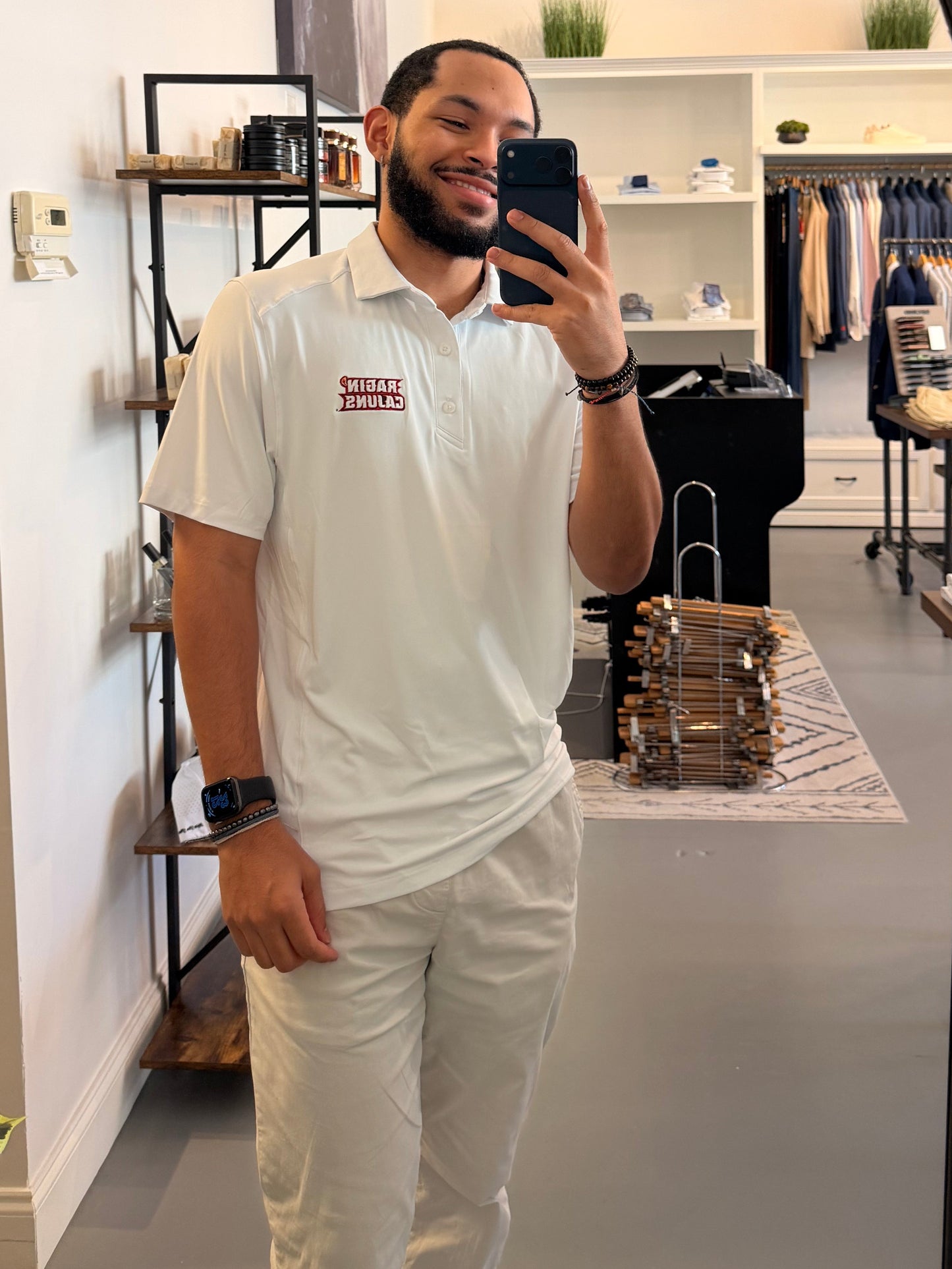 A man takes a mirror selfie in a clothing boutique, wearing a University of Louisiana-Lafayette Ragin Cajuns white polo shirt with a Ragin Cajuns red embroidered logo on the chest and white pants. The boutique has a modern interior with a large arched mirror, clothing racks, and shelves.