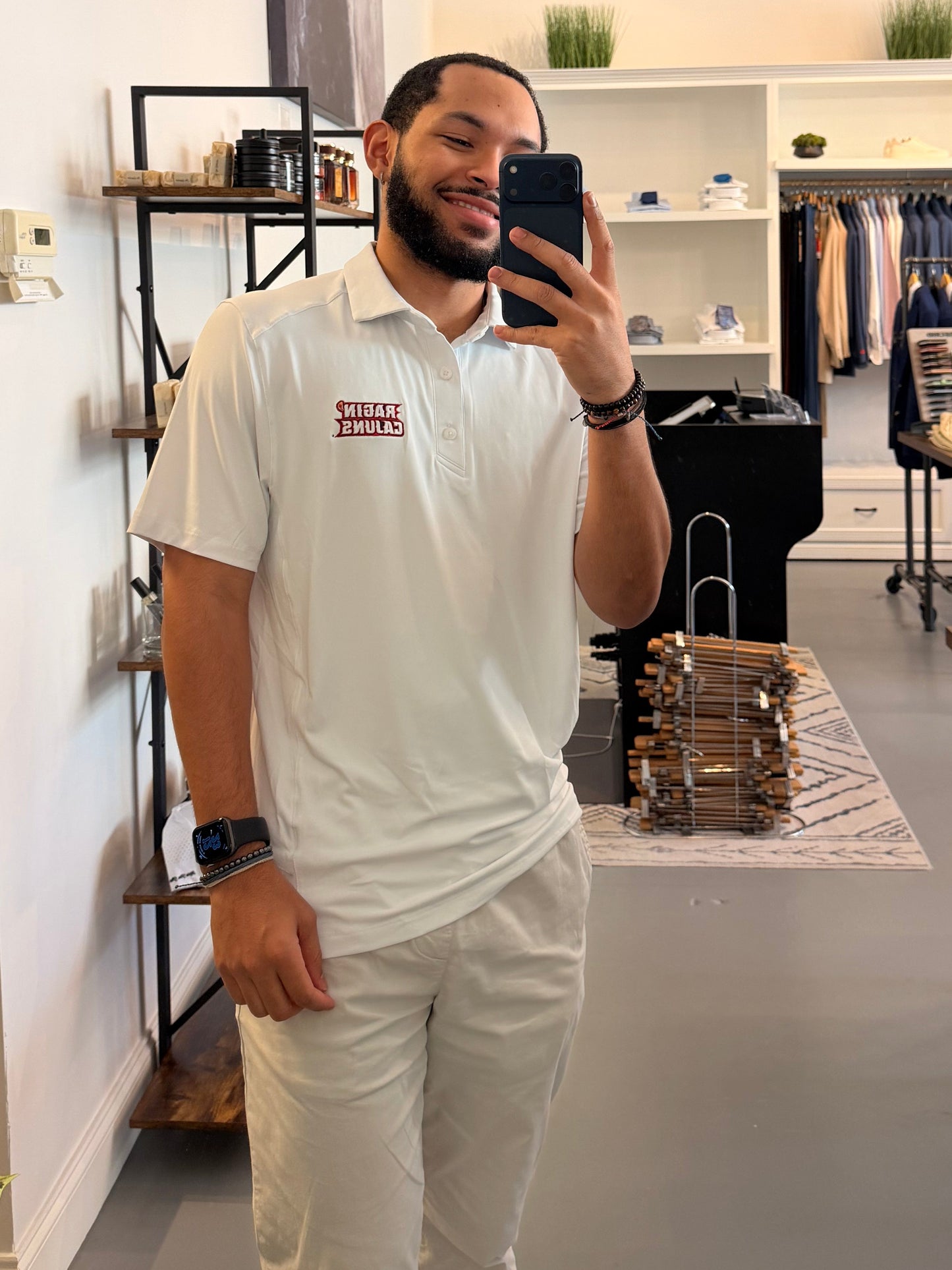 A man takes a mirror selfie in a clothing boutique, wearing a University of Louisiana-Lafayette Ragin Cajuns white polo shirt with a Ragin Cajuns red embroidered logo on the chest and white pants. The boutique has a modern interior with a large arched mirror, clothing racks, and shelves.