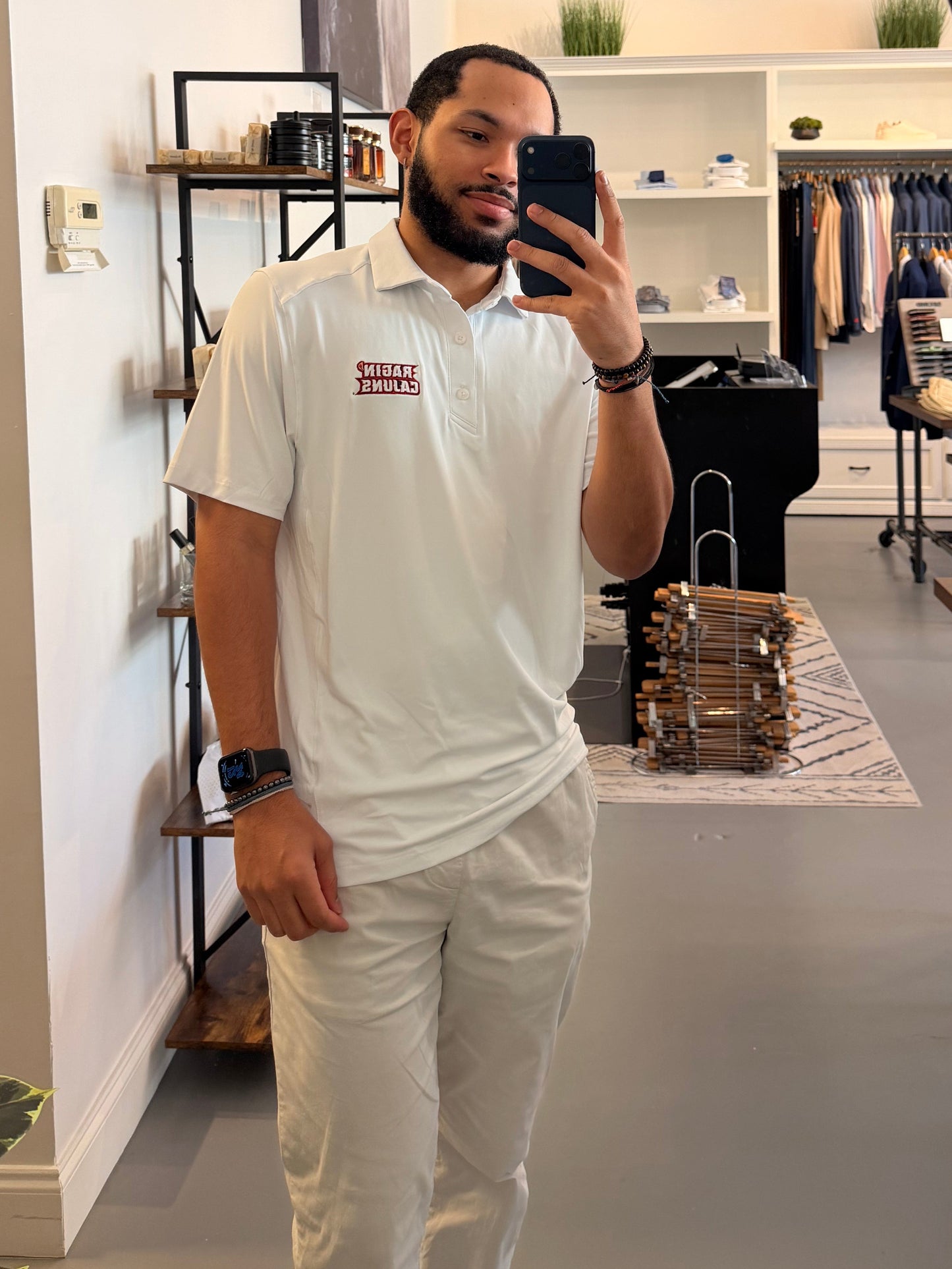 A man takes a mirror selfie in a clothing boutique, wearing a University of Louisiana-Lafayette Ragin Cajuns white polo shirt with a Ragin Cajuns red embroidered logo on the chest and white pants. The boutique has a modern interior with a large arched mirror, clothing racks, and shelves.